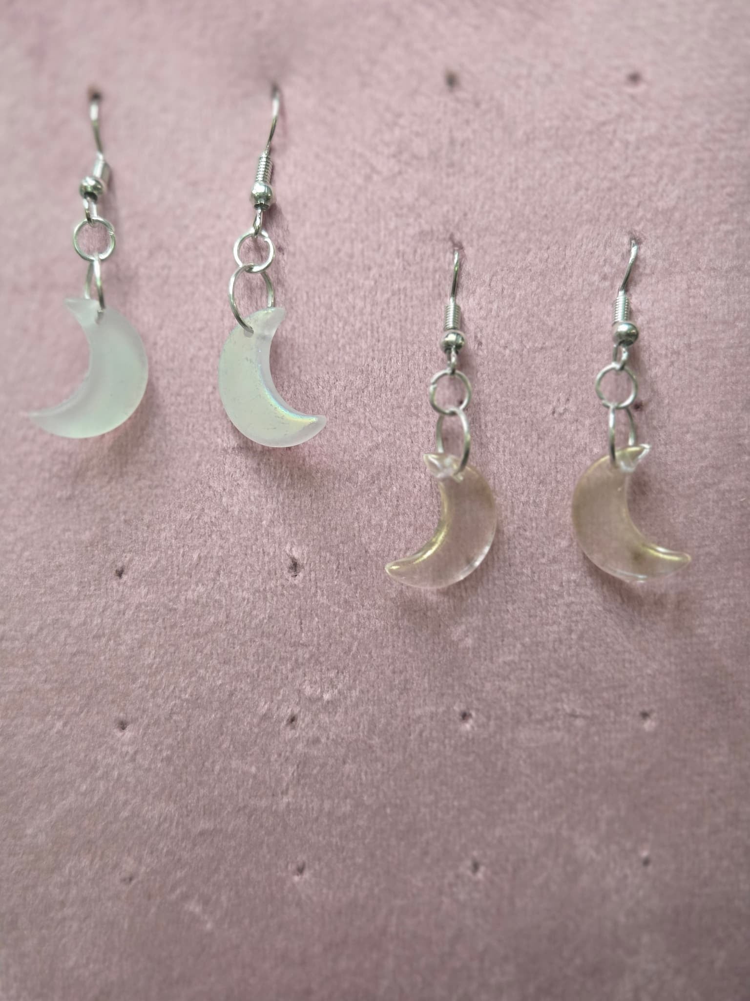 Two pairs of glass crescent moon drop earrings on pink background — frosted white matte finish (left) and clear iridescent AB finish (right), showing size comparison