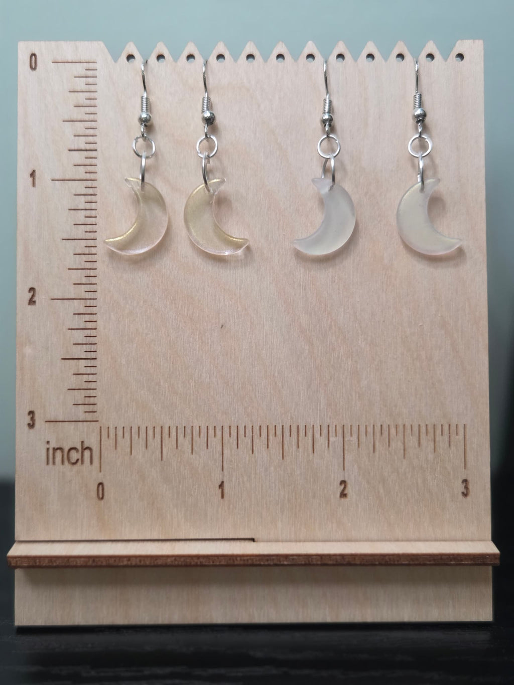 Two pairs of glass crescent moon drop earrings on wooden ruler board showing approximately 1.5 inch length — clear iridescent and frosted white finishes side by side