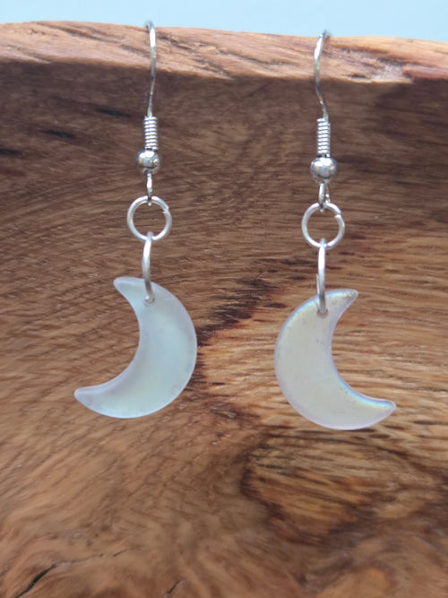Close-up of frosted white matte glass crescent moon drop earrings displayed on rustic wood surface, showing soft cloudy glow