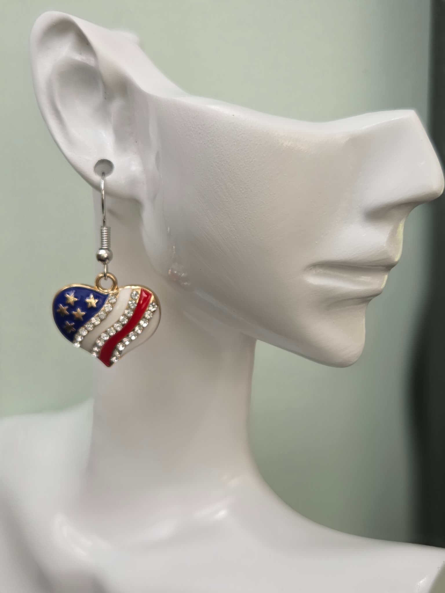 Handmade American flag heart earring on a white mannequin ear, featuring navy blue enamel with gold stars, red and white enamel stripes, and a diagonal clear rhinestone band in a gold-tone setting