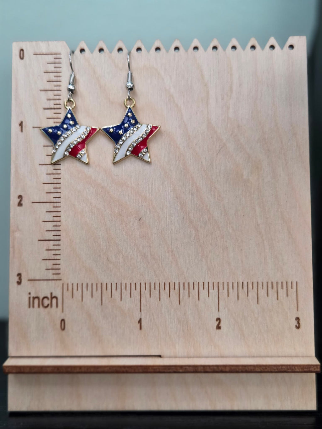 Pair of American flag star dangle earrings on a wooden display board with ruler showing approximately 1 inch charm size, in red, white, and blue enamel with clear rhinestones