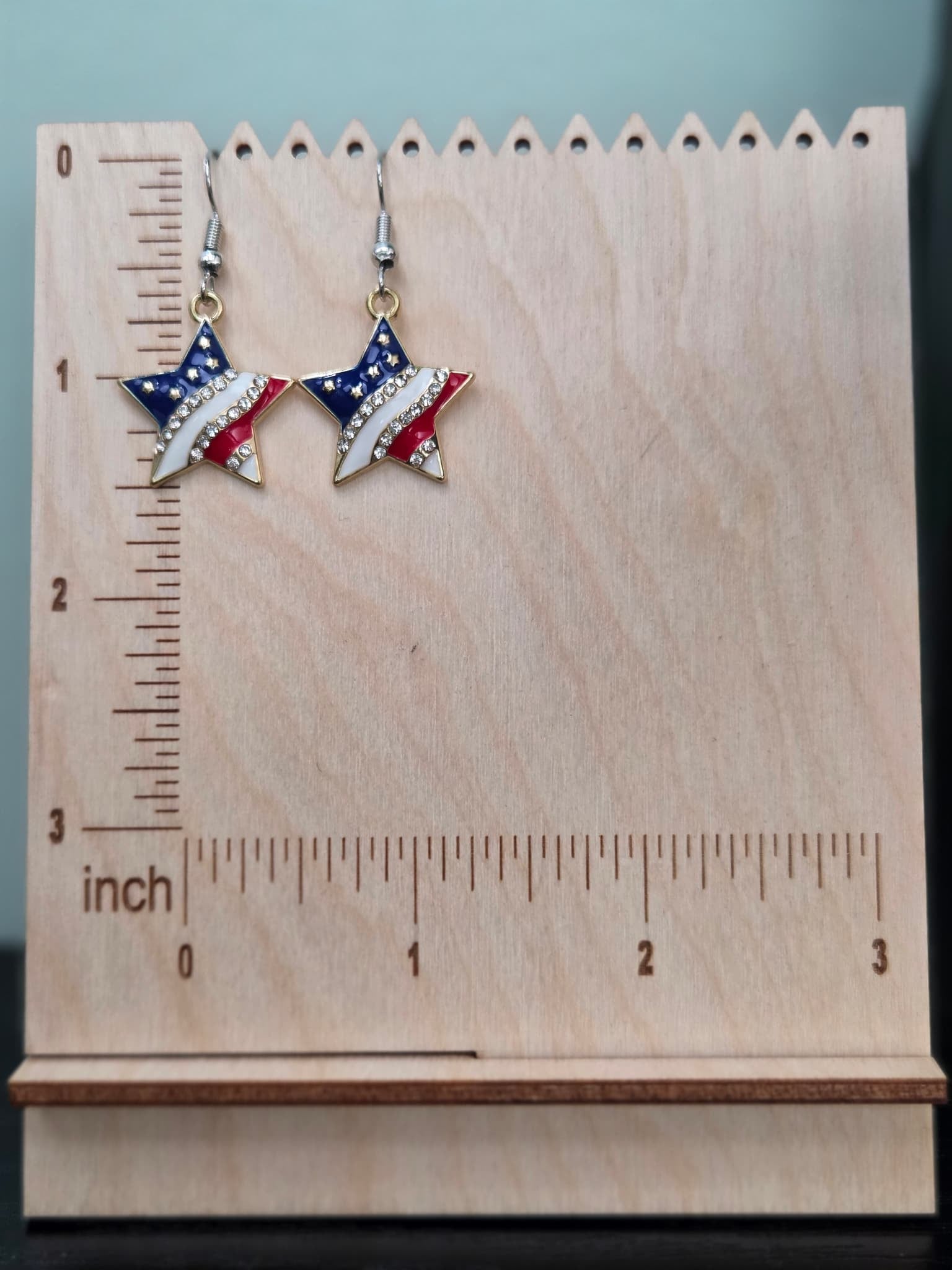Pair of American flag star dangle earrings on a wooden display board with ruler showing approximately 1 inch charm size, in red, white, and blue enamel with clear rhinestones