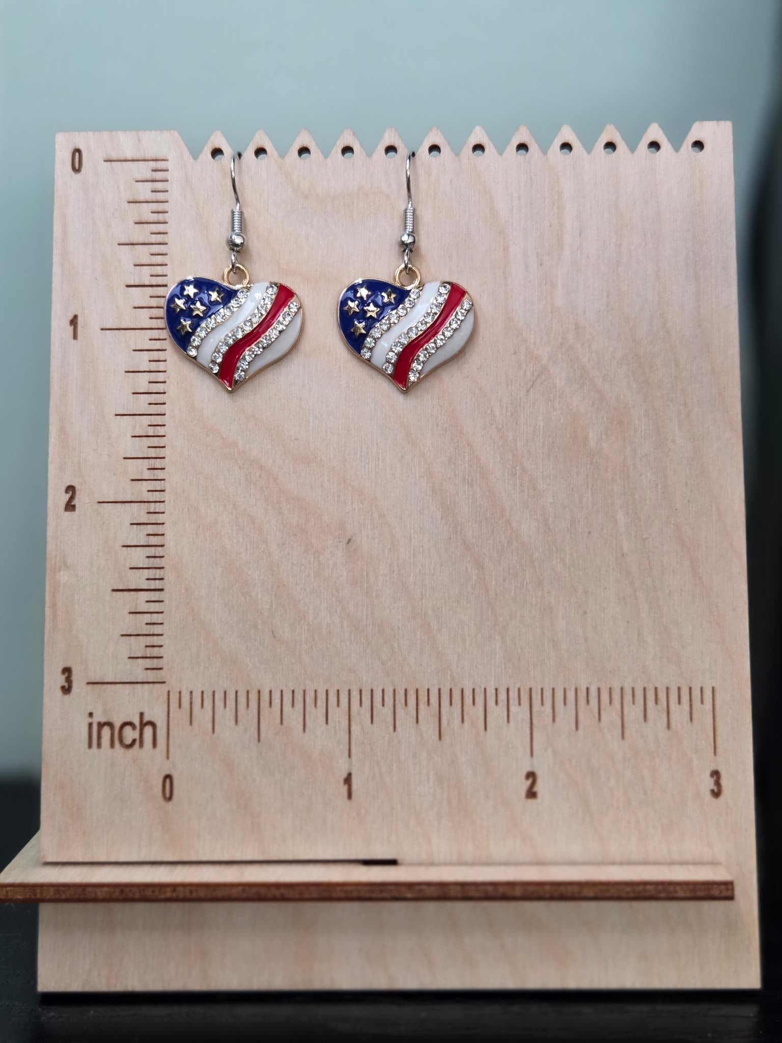 Pair of American flag heart dangle earrings on a wooden display board with ruler showing approximately 1 inch charm size, in red, white, and blue enamel with gold stars and clear rhinestones