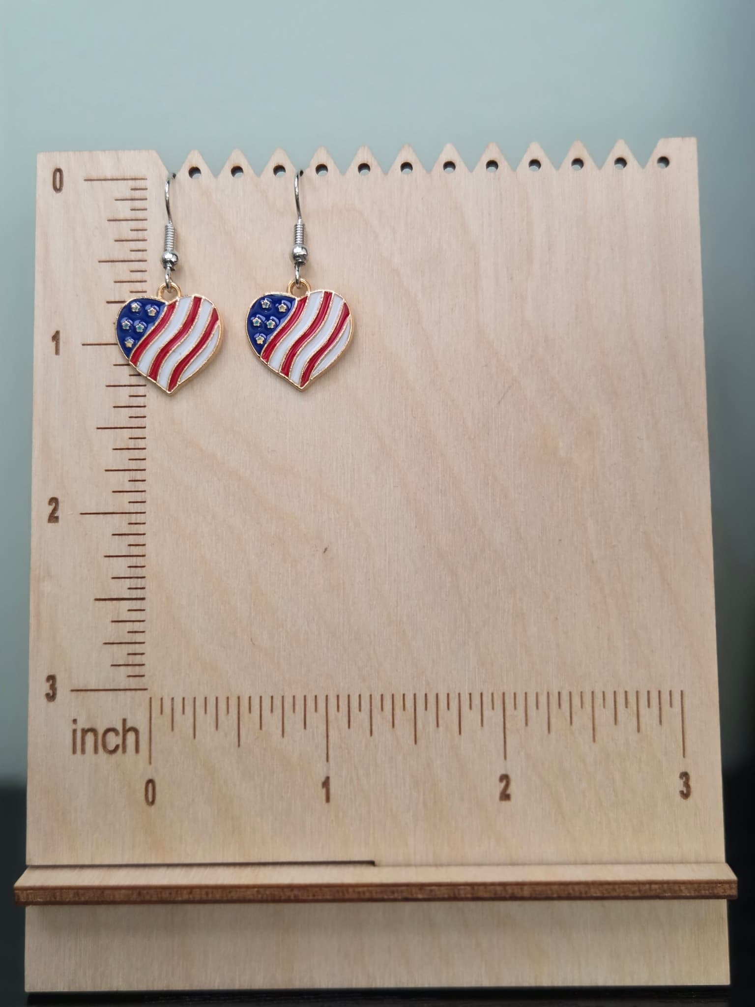 Pair of American flag heart dangle earrings on a wooden display board with ruler showing approximately 1 inch charm size, featuring bold red and white wavy stripes with a navy blue rhinestone star corner in a gold-tone setting