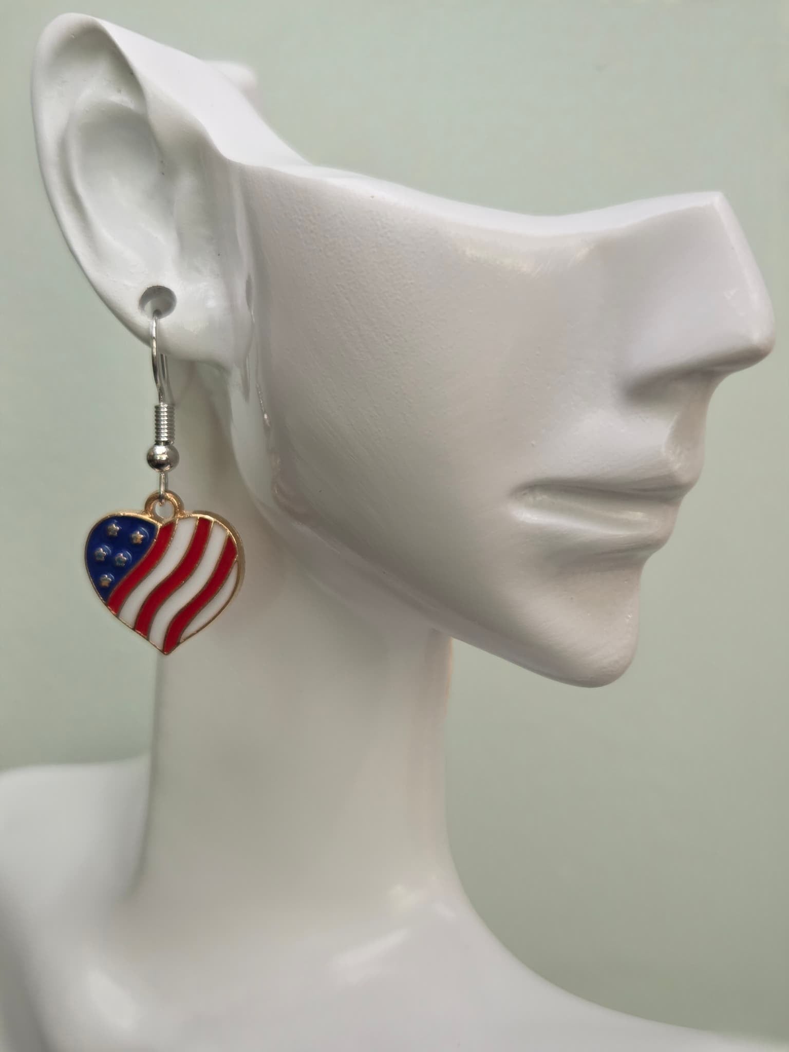 Handmade American flag heart earring on a white mannequin ear, showcasing bold red and white wavy enamel stripes with a navy blue corner and rhinestone stars in a gold-tone setting