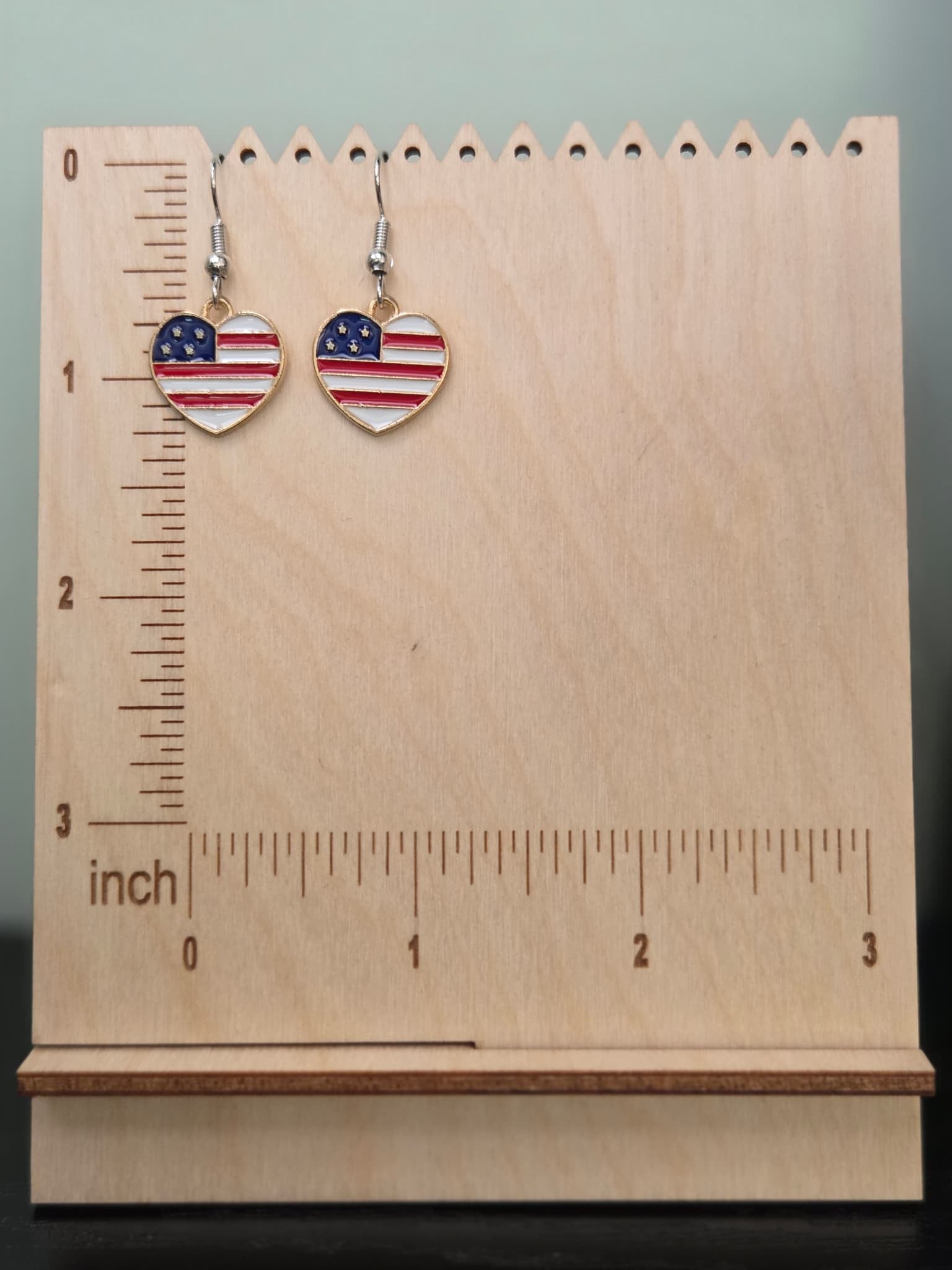 Pair of American flag heart dangle earrings on a wooden display board with ruler showing approximately 1 inch charm size, featuring classic horizontal red and white stripes with a navy blue rhinestone star corner in a gold-tone setting