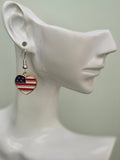 Handmade American flag heart earring on a white mannequin ear, showcasing classic horizontal red and white enamel stripes with a navy blue corner and rhinestone stars in a gold-tone setting