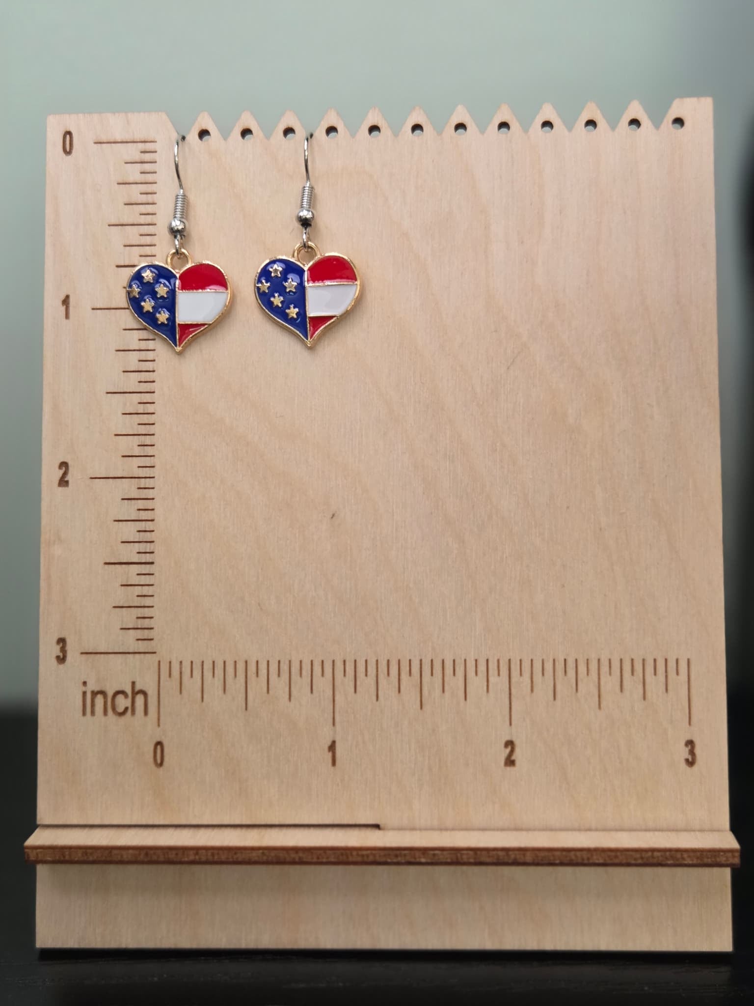 Pair of American flag quartered heart dangle earrings on a wooden display board with ruler showing approximately 1 inch charm size, featuring alternating navy blue with gold stars and red with white stripe sections in a gold-tone setting