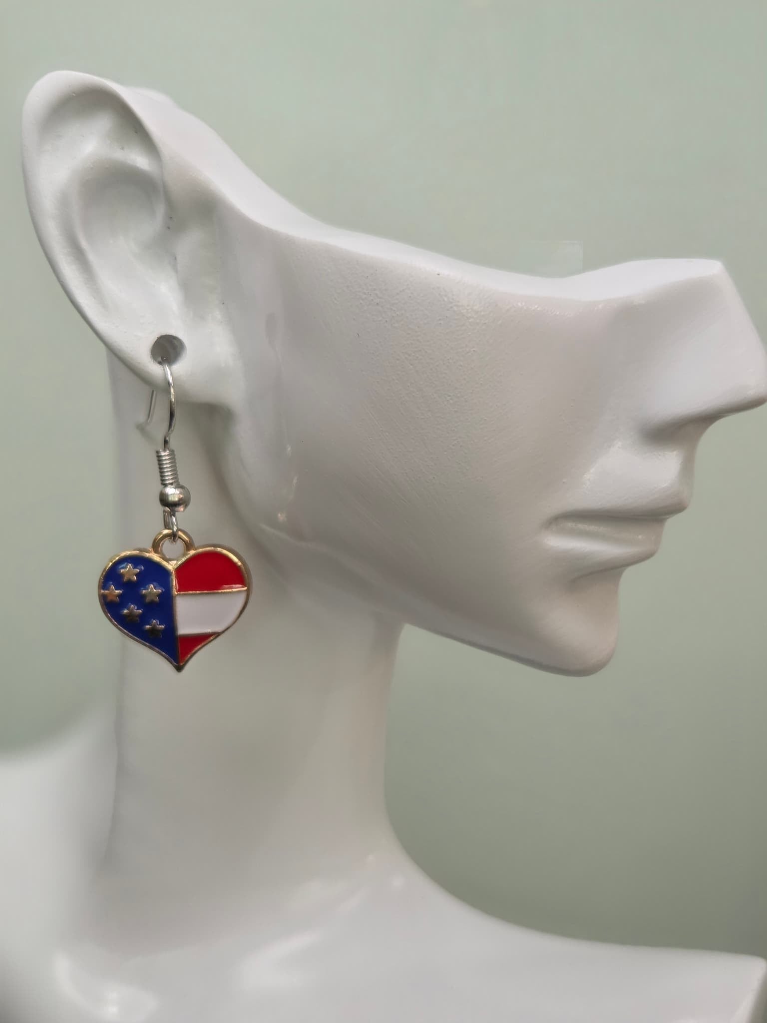 Handmade American flag quartered heart earring on a white mannequin ear, showcasing alternating navy blue enamel with gold stars and red with white stripe enamel sections in a gold-tone setting