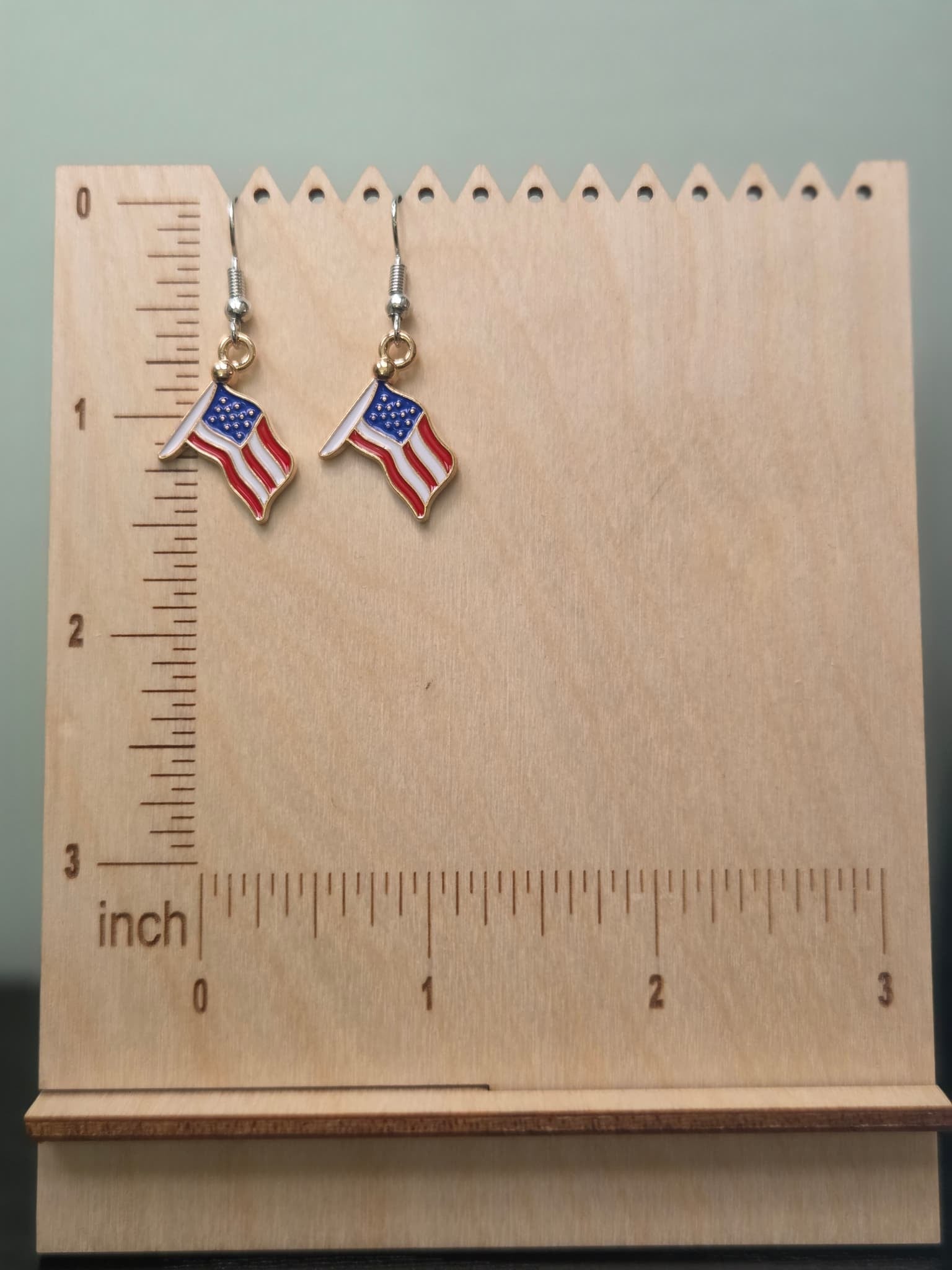 Pair of waving American flag dangle earrings on a wooden display board with ruler showing approximately ¾ inch charm size, featuring red and white wavy stripes with a navy blue star field in a gold-tone frame