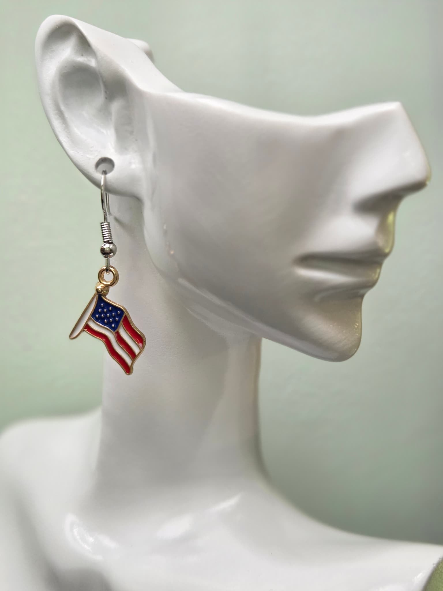 Handmade waving American flag earring on a white mannequin ear, showcasing red and white wavy enamel stripes with a navy blue star field in a gold-tone frame