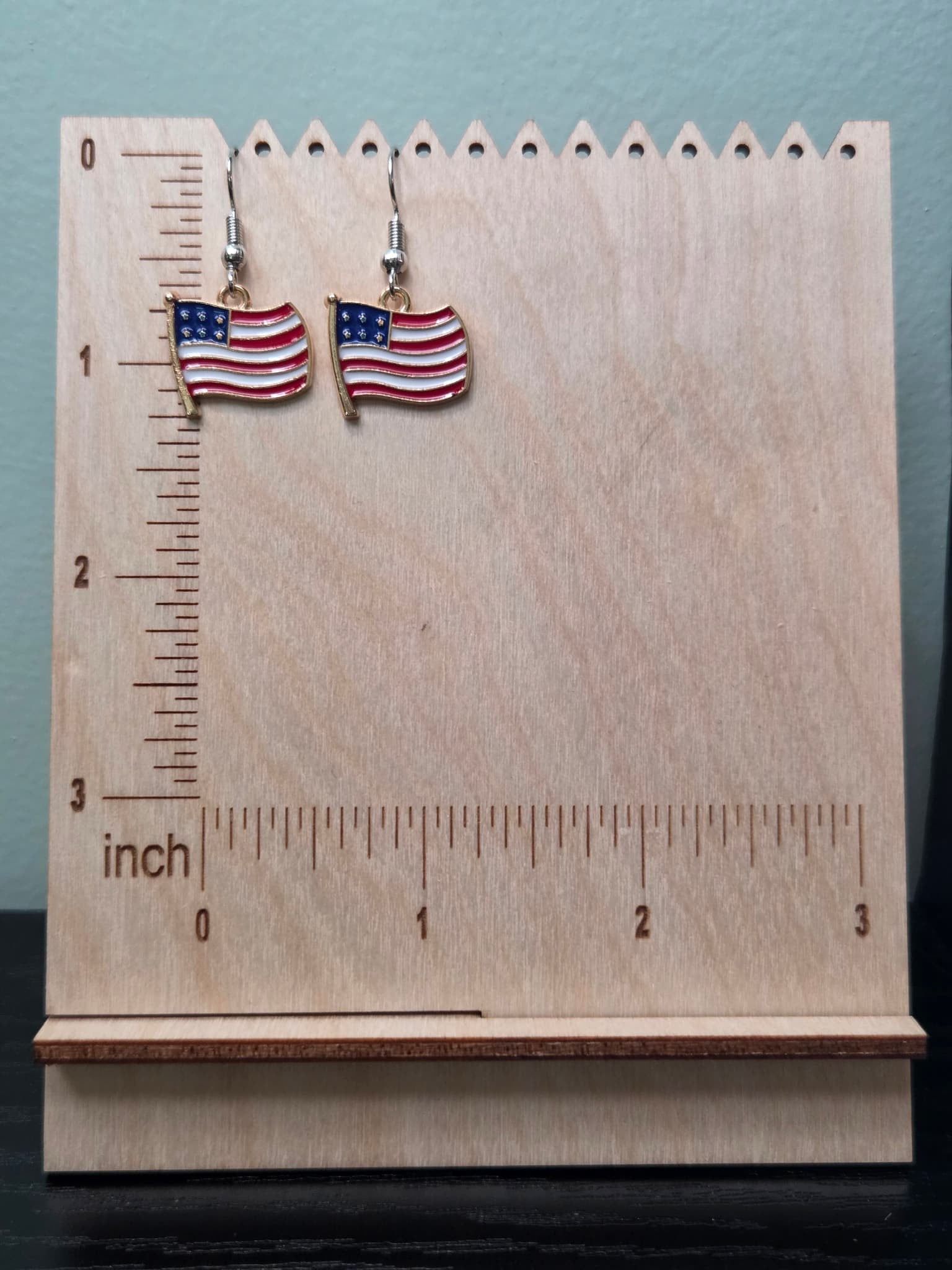 Pair of waving American flag dangle earrings with gold flagpole on a wooden display board with ruler showing approximately 1 inch charm size, featuring red and white wavy stripes with a navy blue star field and cream border in a gold-tone frame