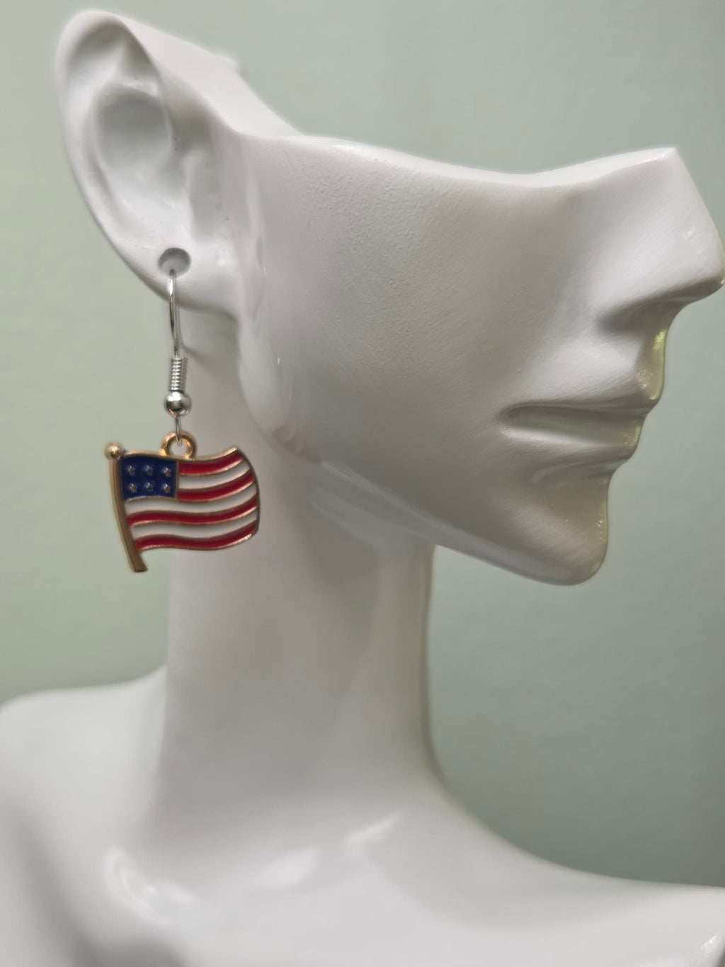 Handmade waving American flag earring with gold flagpole on a white mannequin ear, showcasing red and white wavy enamel stripes with a navy blue star field and cream-toned border