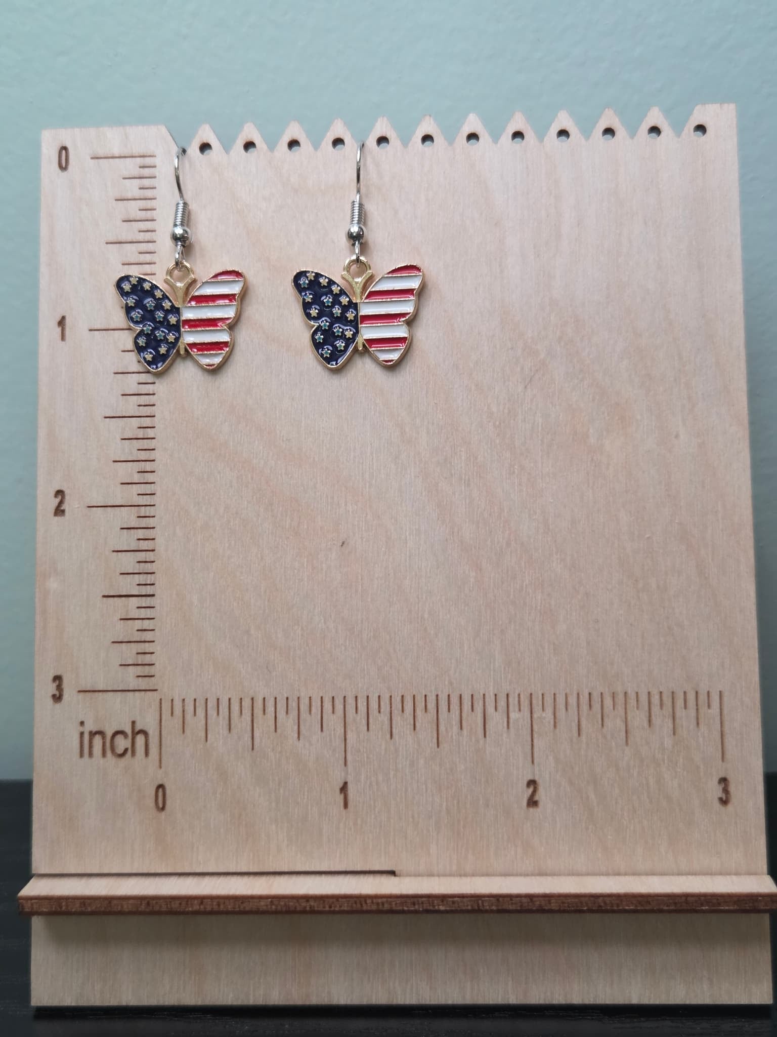 Pair of American flag butterfly dangle earrings on a wooden display board with ruler showing approximately 1 inch charm size, featuring navy blue star wings and red and white striped wings in a gold-tone frame