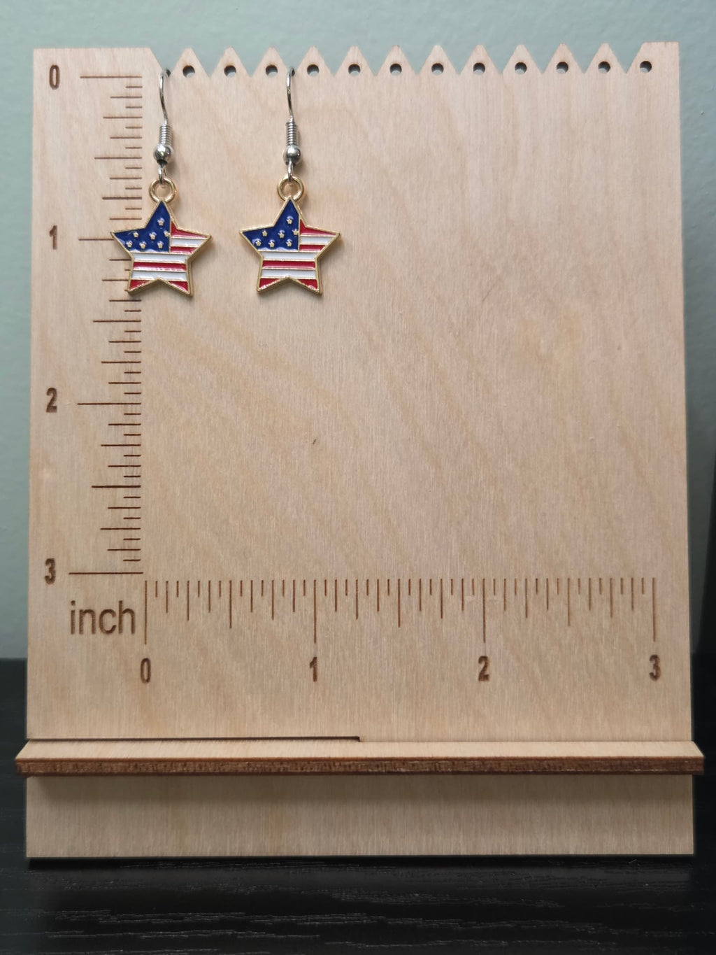 Pair of American flag star dangle earrings on a wooden display board with ruler showing approximately ¾ inch charm size, featuring navy blue corner with white stars and bold red and white stripes in a gold-tone frame