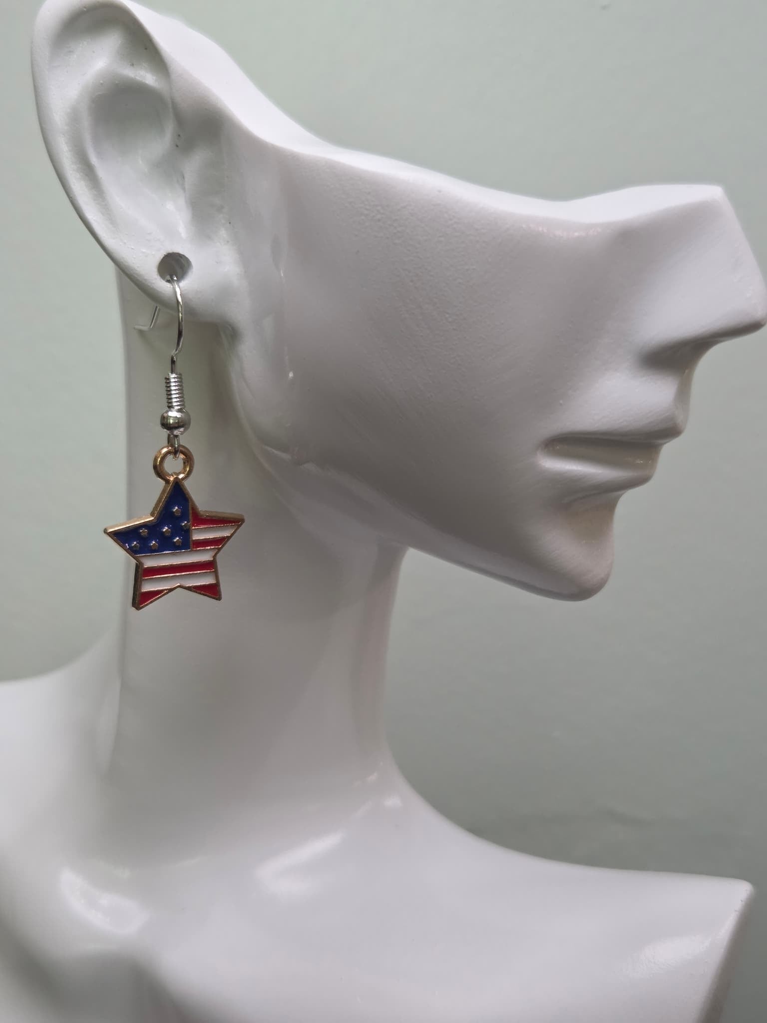 Handmade American flag star earring on a white mannequin ear, showcasing a star-shaped charm with navy blue corner, white stars, and bold red and white enamel stripes in a gold-tone frame