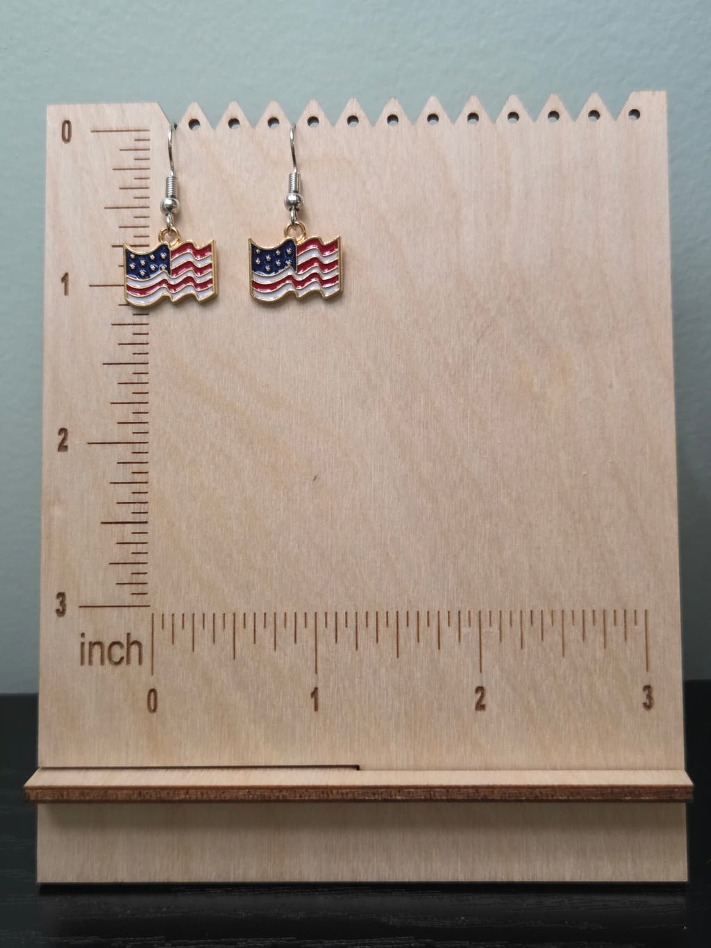 Pair of petite waving American flag dangle earrings on a wooden display board with ruler showing approximately ¾ inch charm size, featuring red and white wavy stripes with a navy blue star corner and gold-tone edge