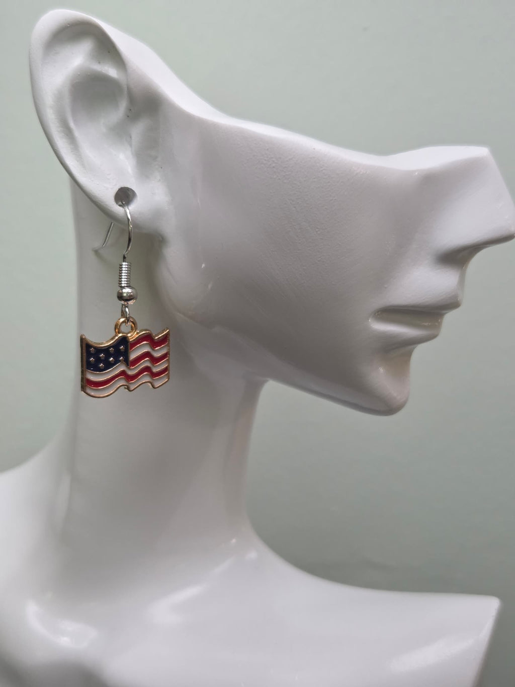 Pair of petite waving American flag dangle earrings on a wooden display board with ruler showing approximately ¾ inch charm size, featuring red and white wavy stripes with a navy blue star corner and gold-tone edge