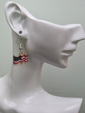 Pair of petite waving American flag dangle earrings on a wooden display board with ruler showing approximately ¾ inch charm size, featuring red and white wavy stripes with a navy blue star corner and gold-tone edge