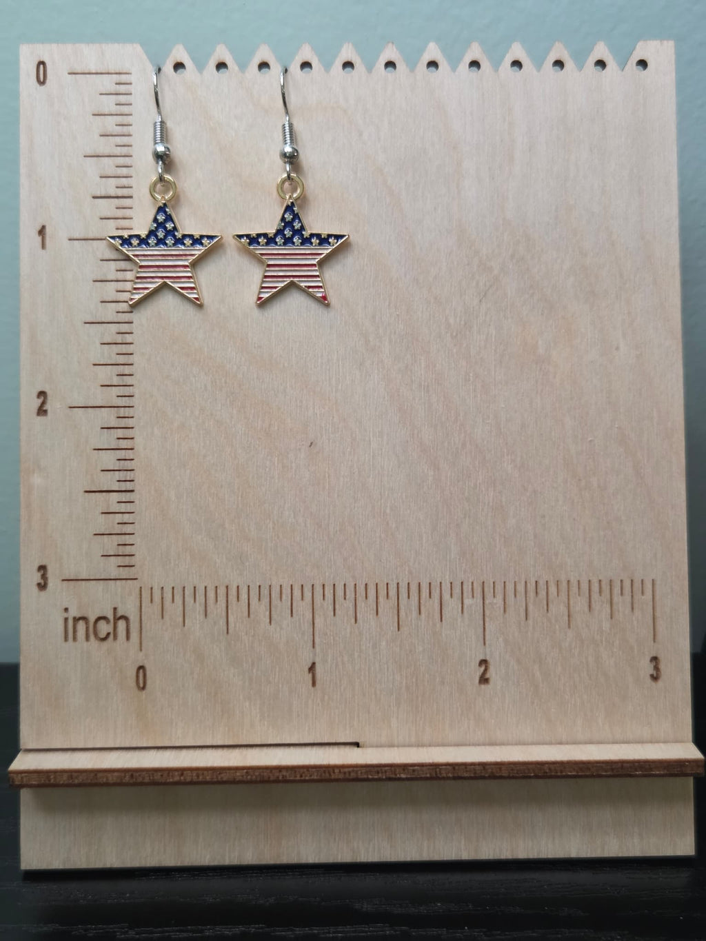 Pair of American flag bold star dangle earrings on a wooden display board with ruler showing approximately ¾ inch charm size, featuring wide navy blue upper section with white stars and bold red and white horizontal stripes in a gold-tone frame
