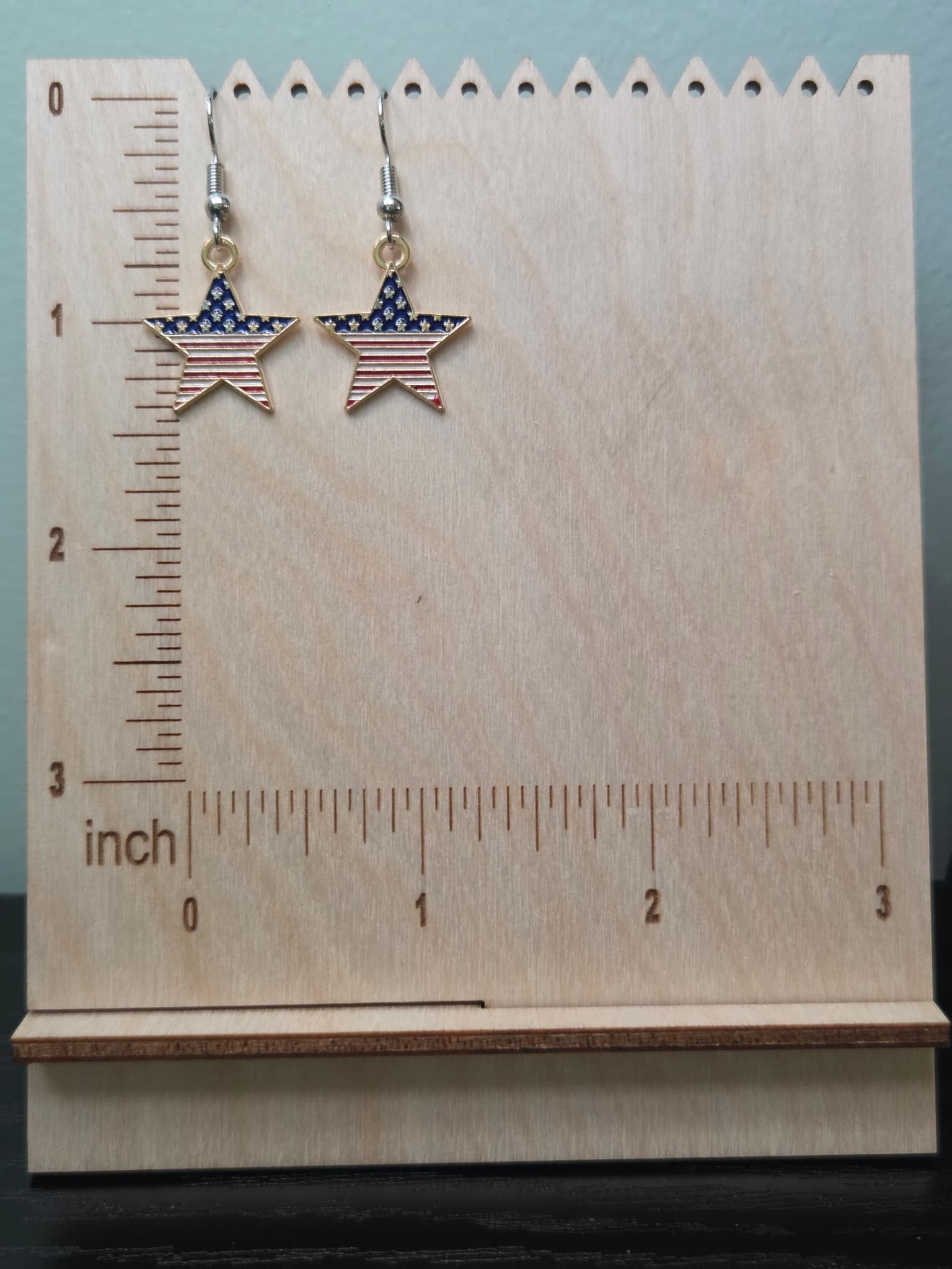 Pair of American flag bold star dangle earrings on a wooden display board with ruler showing approximately ¾ inch charm size, featuring wide navy blue upper section with white stars and bold red and white horizontal stripes in a gold-tone frame