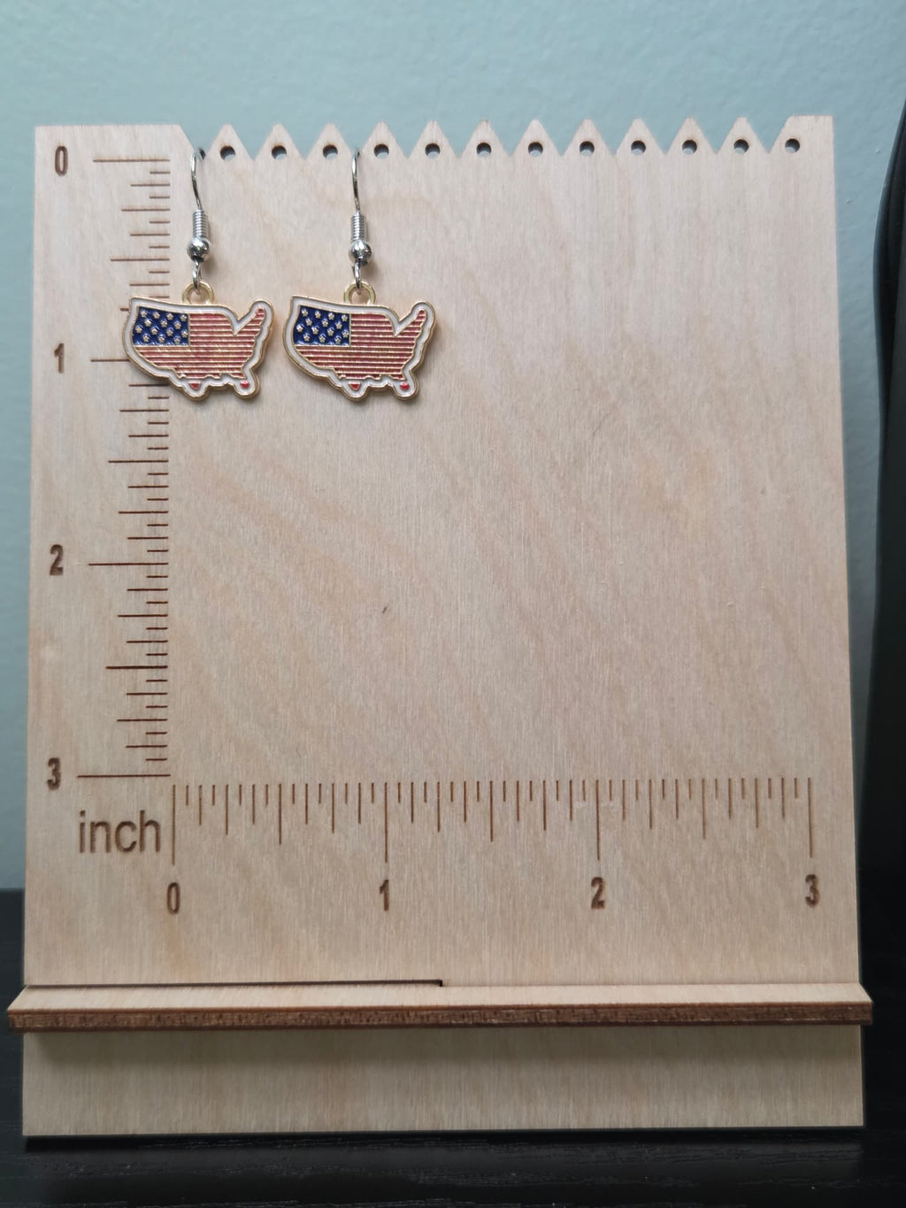 Pair of USA map-shaped dangle earrings on a wooden display board with ruler showing approximately ¾ inch charm size, filled with American flag design featuring navy blue star corner and bold red and white horizontal stripes in a gold-tone edge
