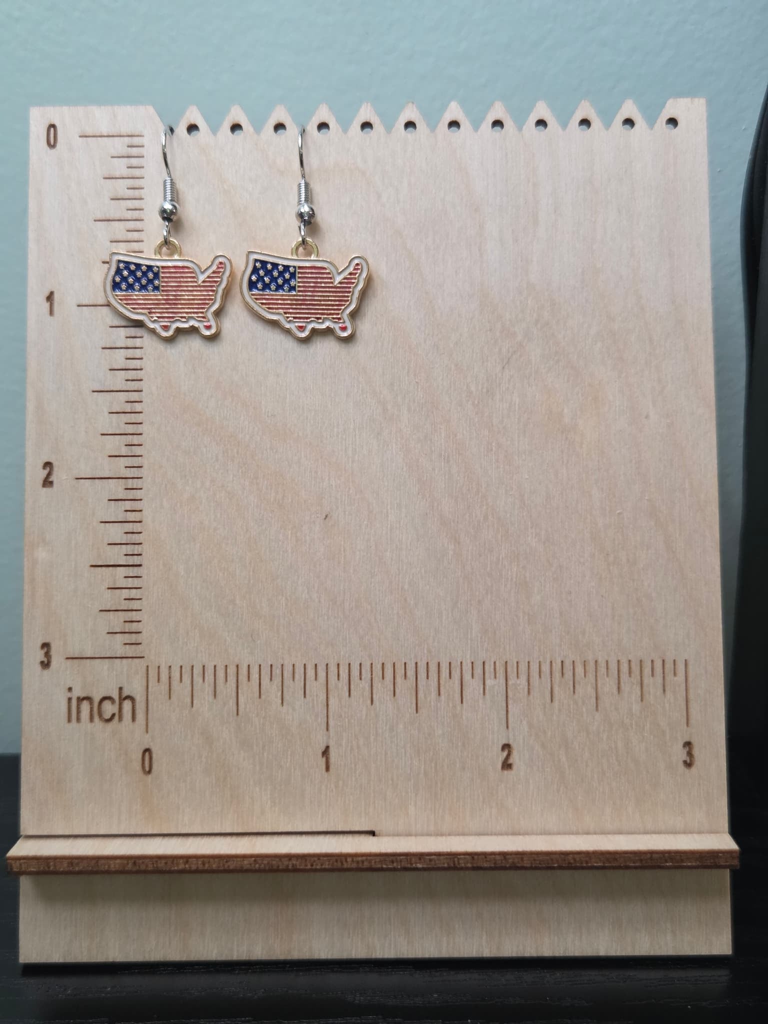 Pair of USA map-shaped dangle earrings on a wooden display board with ruler showing approximately ¾ inch charm size, filled with American flag design featuring navy blue star corner and bold red and white horizontal stripes in a gold-tone edge