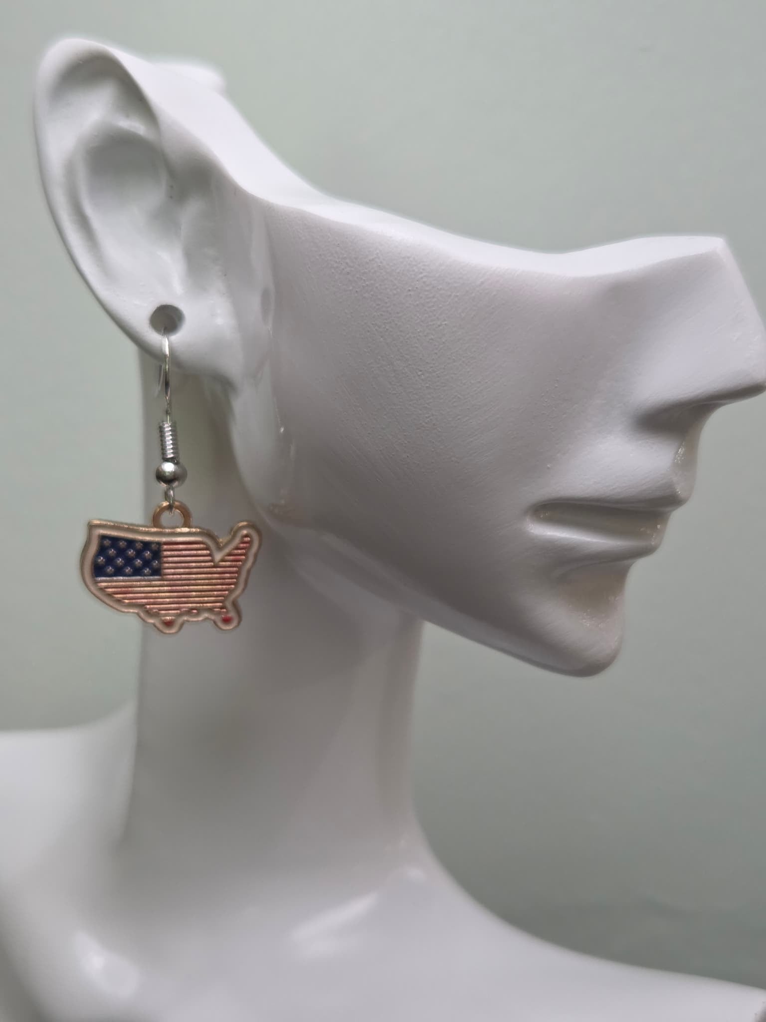 Handmade USA map-shaped earring on a white mannequin ear, featuring a continental United States silhouette filled with navy blue star corner and bold red and white enamel stripes in a gold-tone edge