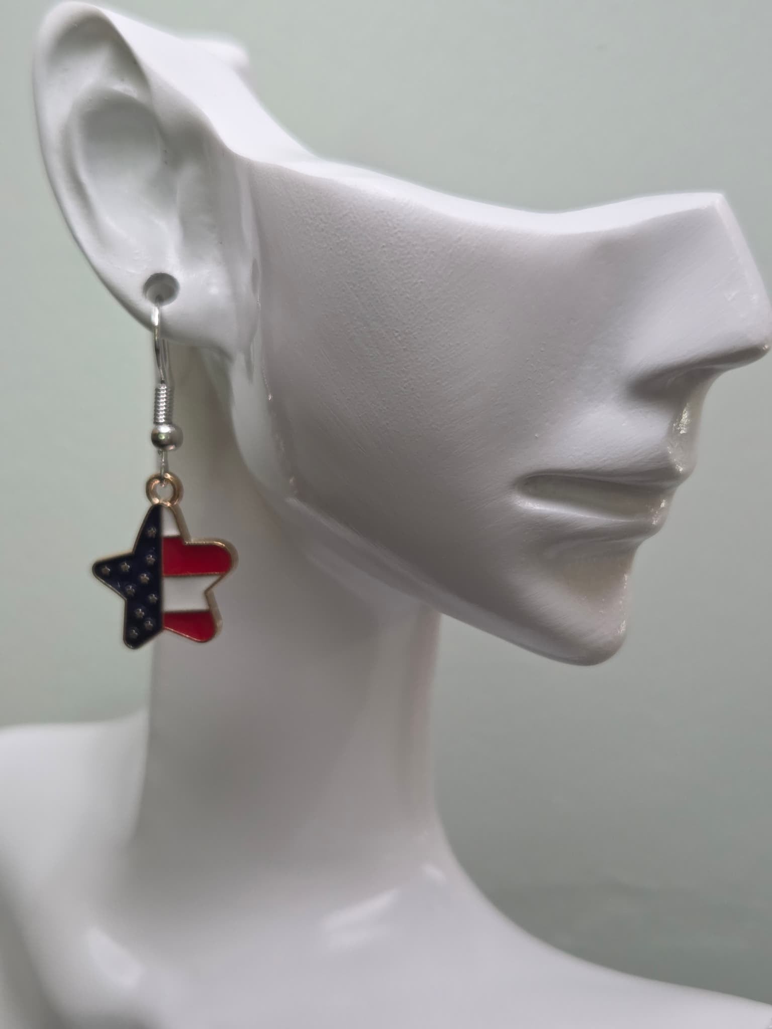 Handmade American flag diagonal star earring on a white mannequin ear, featuring a star-shaped charm with navy blue enamel and white stars on the upper half and bold red and white diagonal stripes on the lower half in a gold-tone frame