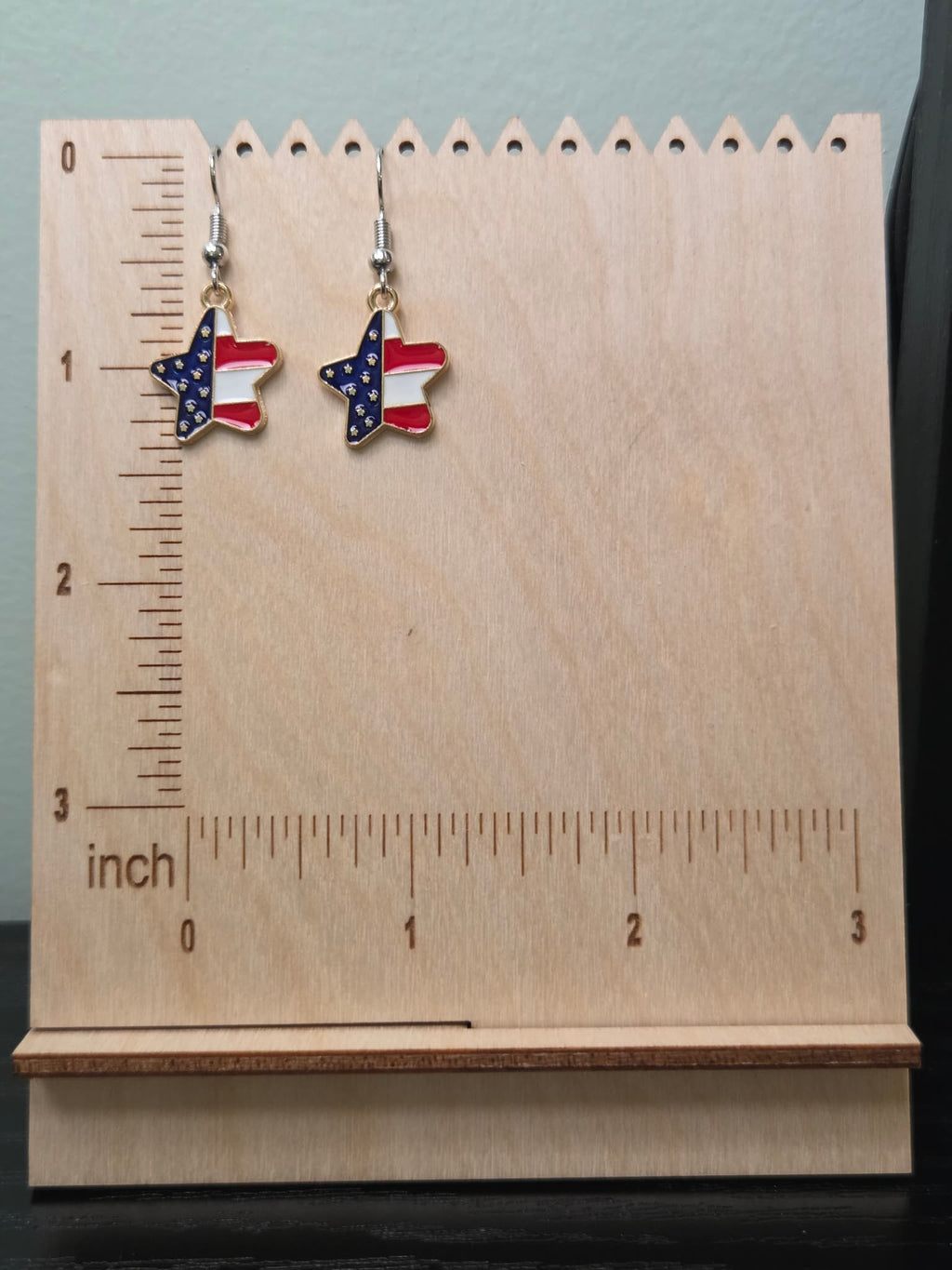 Pair of American flag diagonal star dangle earrings on a wooden display board with ruler showing approximately ¾ inch charm size, featuring navy blue star upper half and red and white stripe lower half in a gold-tone frame