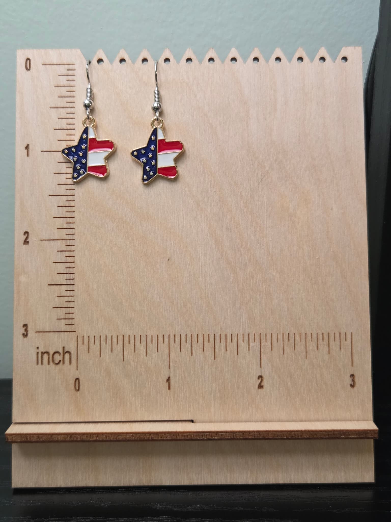 Pair of American flag diagonal star dangle earrings on a wooden display board with ruler showing approximately ¾ inch charm size, featuring navy blue star upper half and red and white stripe lower half in a gold-tone frame
