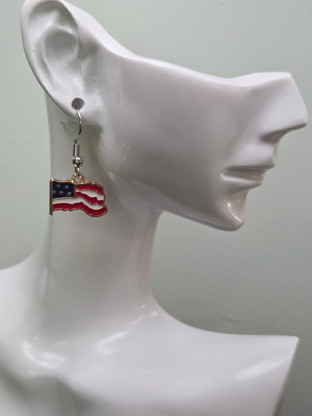 Handmade waving American flag earring on a white mannequin ear, featuring bold rippled red and white enamel stripes with a navy blue corner of white stars and a gold-tone edge