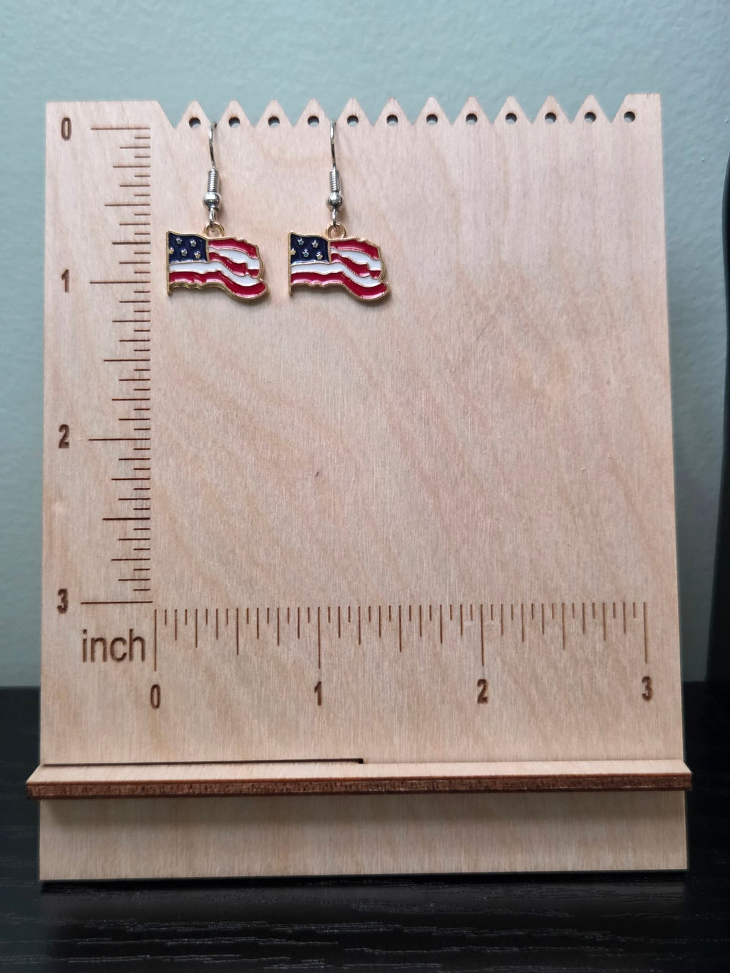  Pair of waving American flag dangle earrings on a wooden display board with ruler showing approximately ¾ inch charm size, featuring bold rippled red and white stripes with a navy blue star corner and gold-tone edge