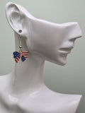 Handmade American flag bow earring on a white mannequin ear, featuring a bow-shaped charm with navy blue enamel and white stars on the left wing and bold red and white stripes on the right wing in a gold-tone frame