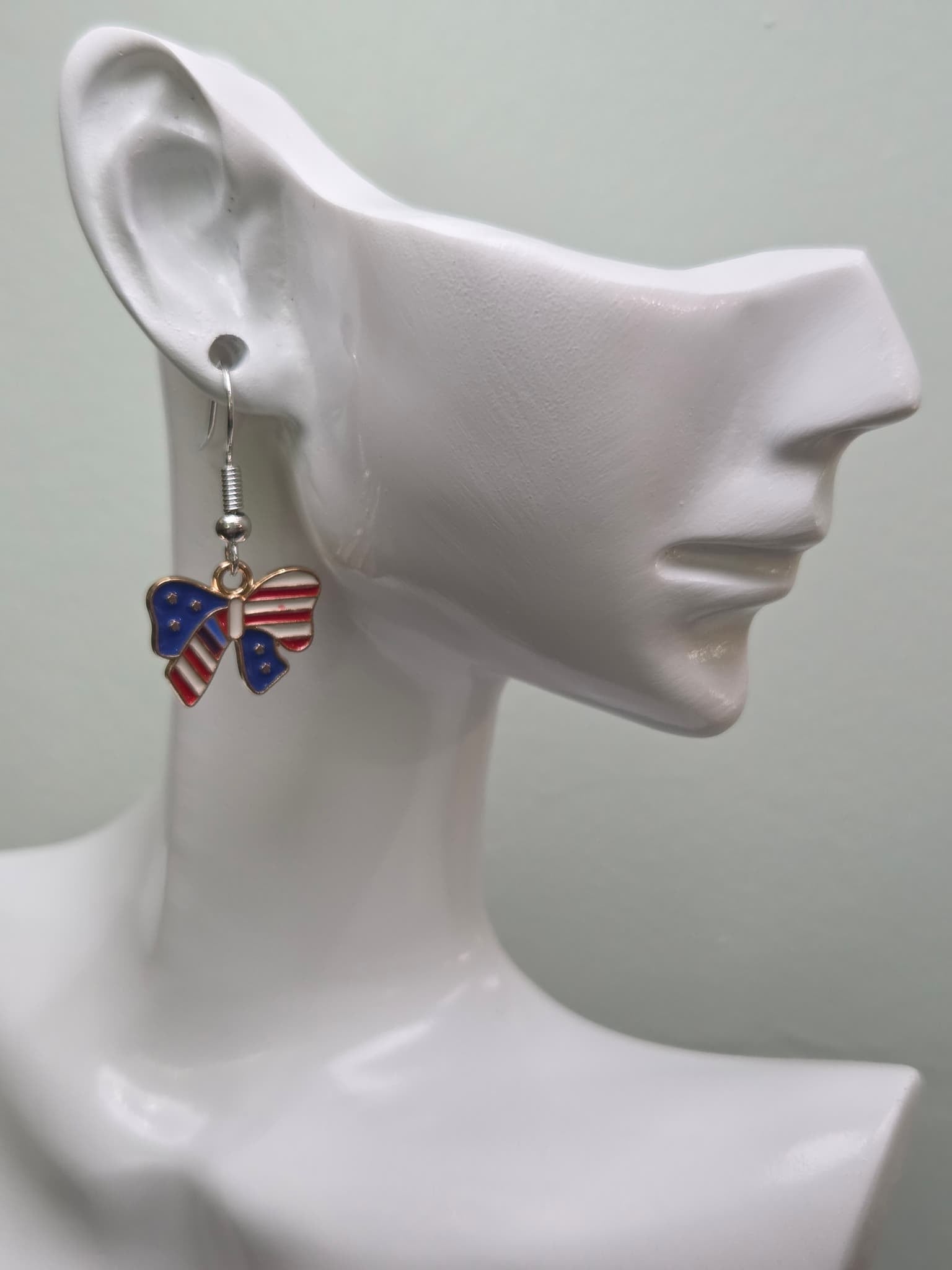 Handmade American flag bow earring on a white mannequin ear, featuring a bow-shaped charm with navy blue enamel and white stars on the left wing and bold red and white stripes on the right wing in a gold-tone frame