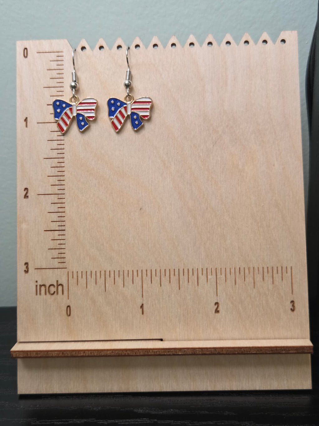 Pair of American flag bow dangle earrings on a wooden display board with ruler showing approximately ¾ inch charm size, featuring navy blue star left wing and red and white stripe right wing in a gold-tone frame