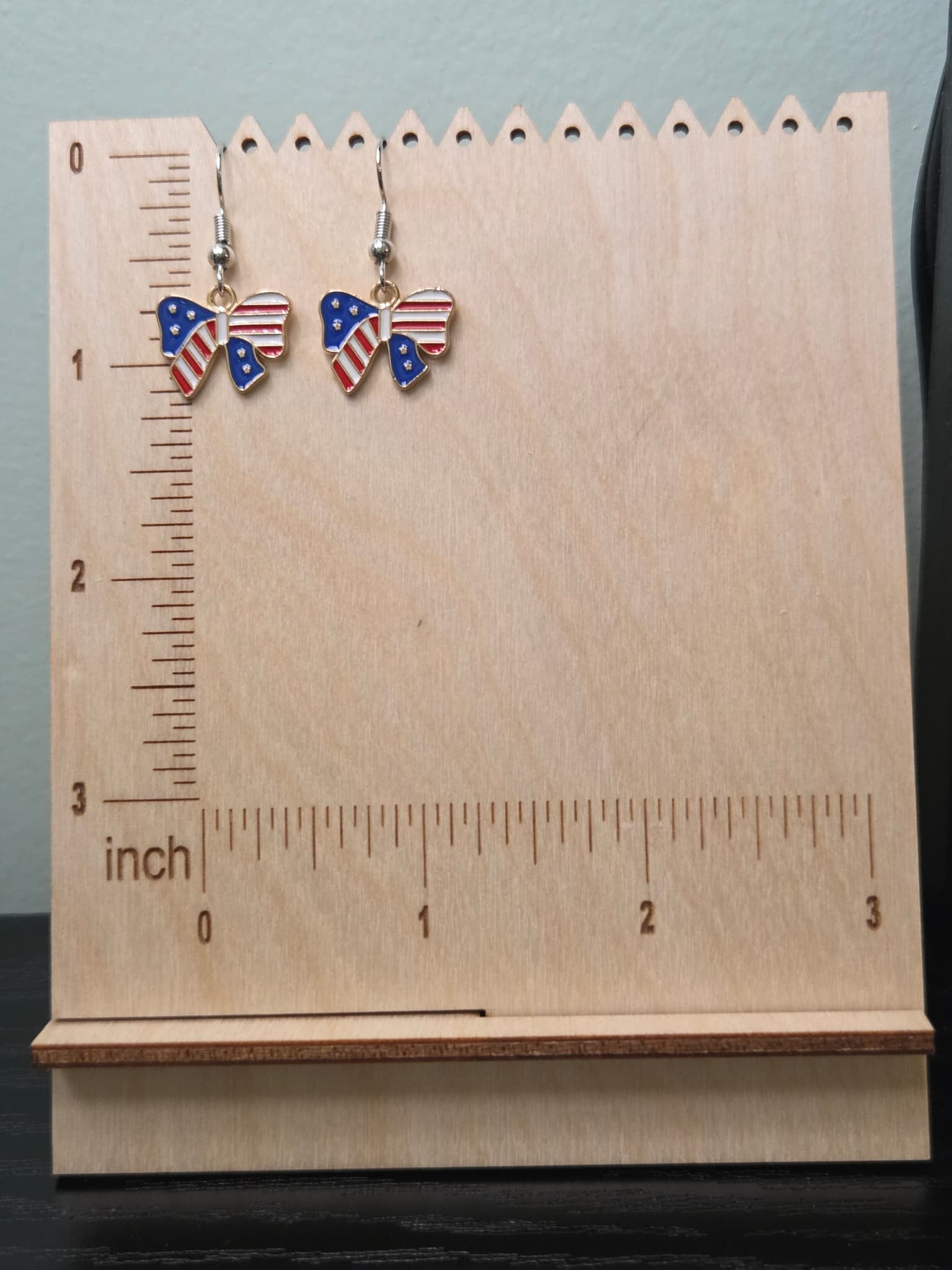 Pair of American flag bow dangle earrings on a wooden display board with ruler showing approximately ¾ inch charm size, featuring navy blue star left wing and red and white stripe right wing in a gold-tone frame