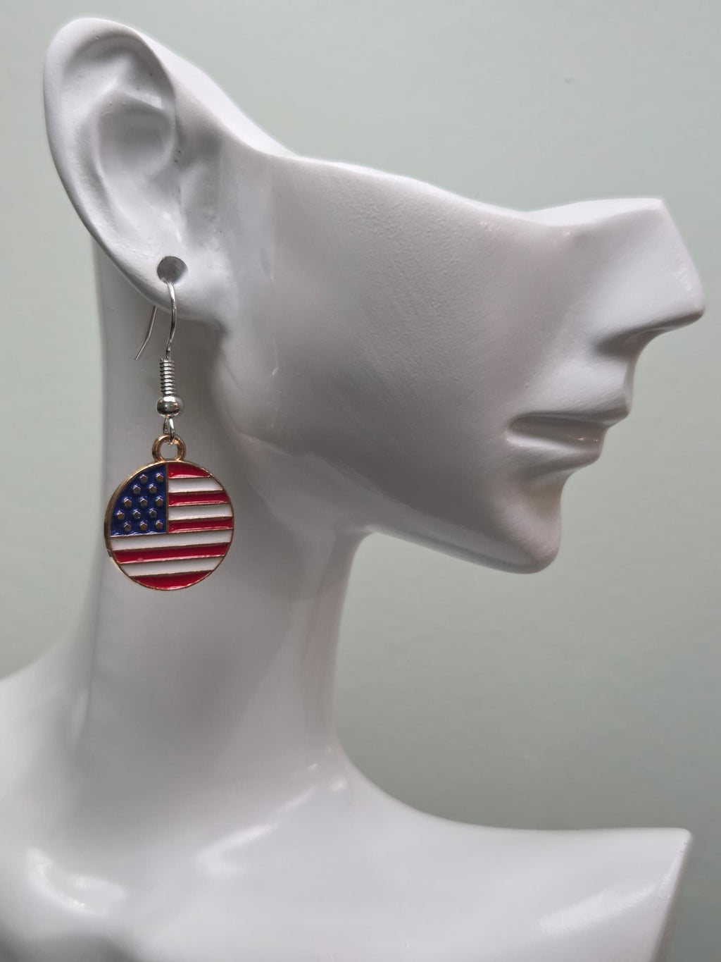 Handmade round American flag earring on a white mannequin ear, featuring a circular gold-tone bordered charm with bold red and white stripes and a navy blue corner of white stars