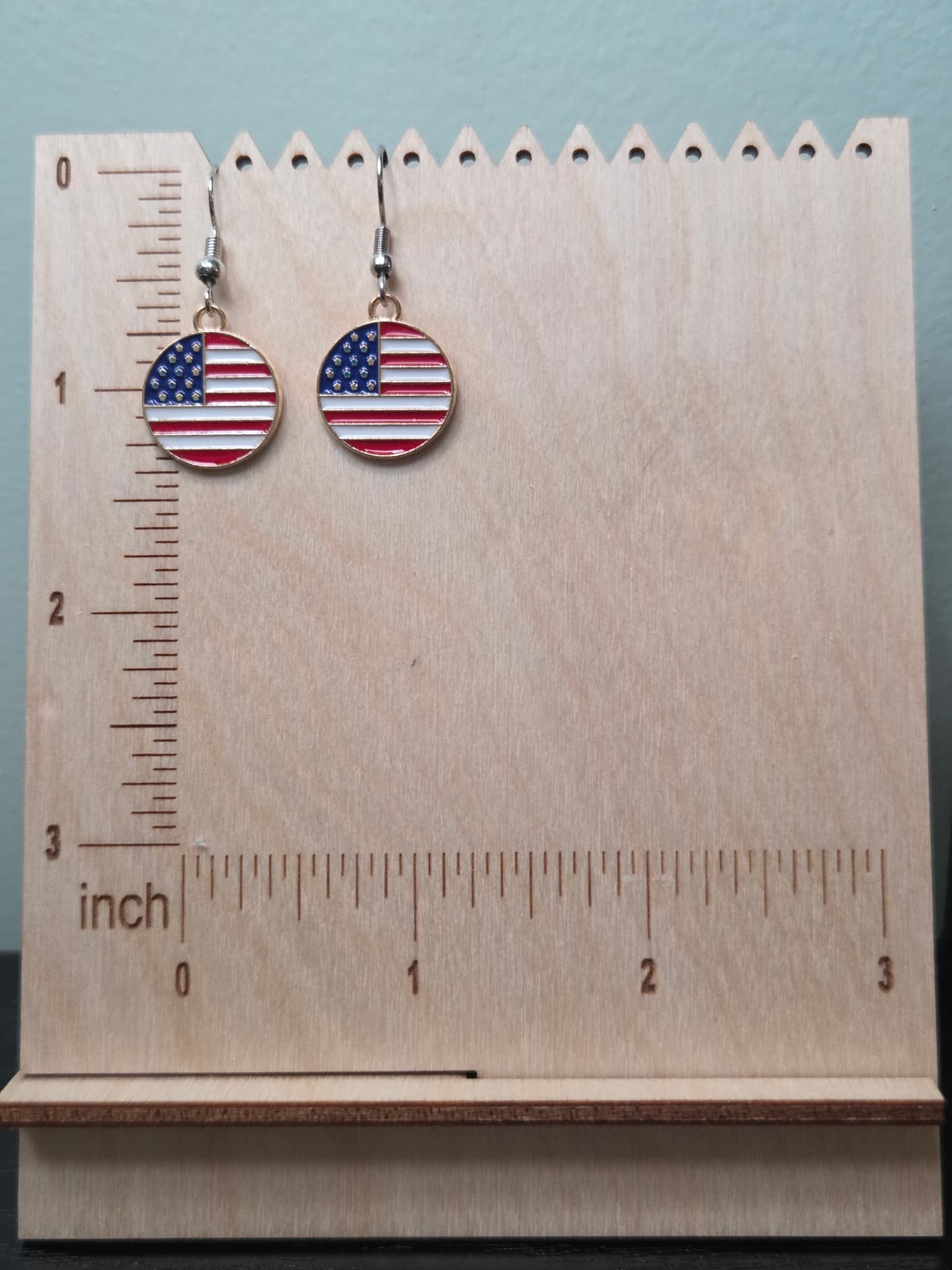 Pair of round American flag dangle earrings on a wooden display board with ruler showing approximately ¾ inch charm size, featuring red and white stripes with a navy blue star corner in a gold-tone circular border