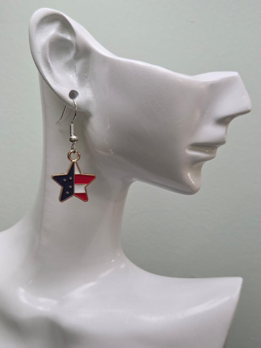 Handmade petite American flag split star earring on a white mannequin ear, featuring a star-shaped charm with navy blue enamel and white stars on the upper left and vivid red and white diagonal stripes on the lower right in a gold-tone frame