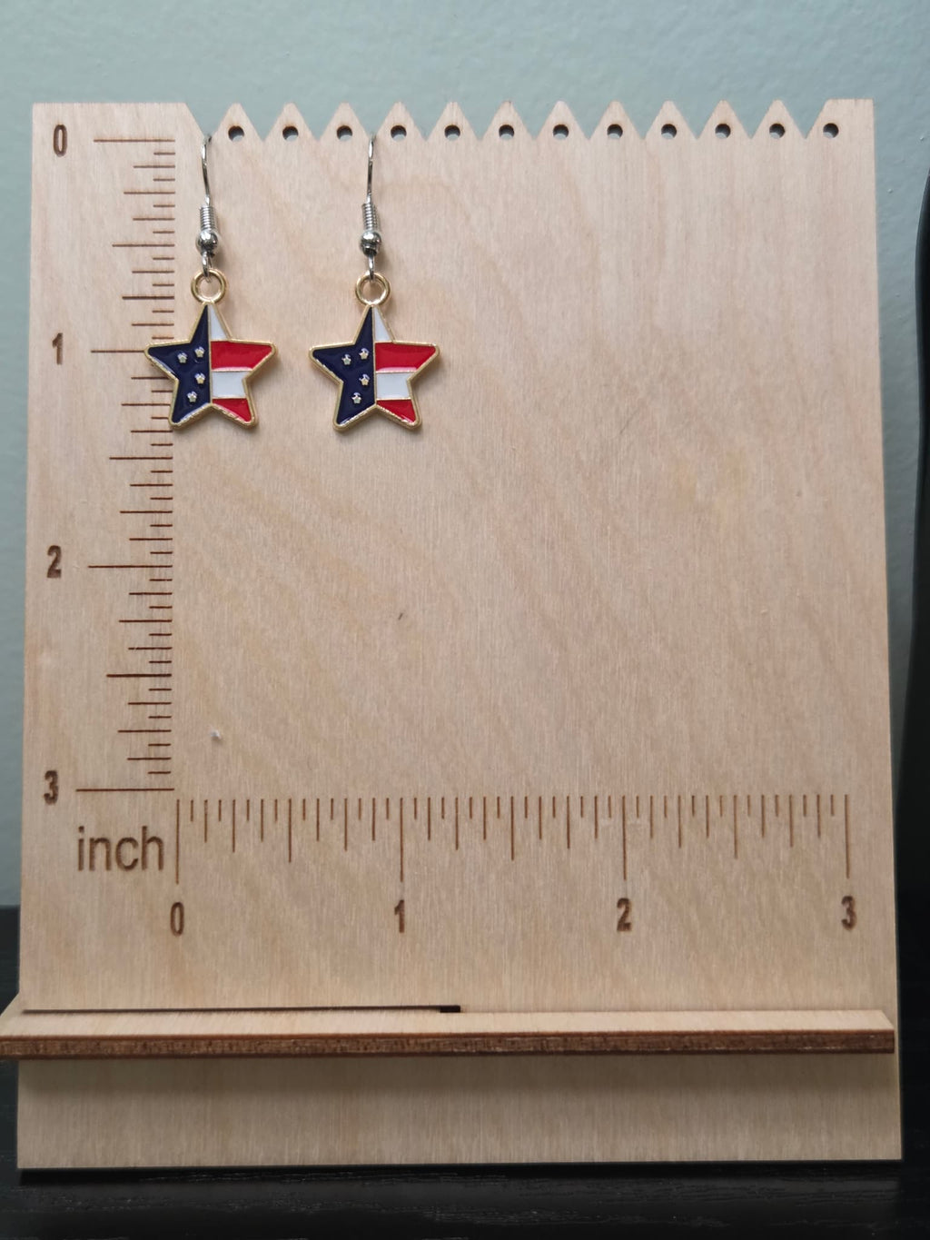 Pair of petite American flag split star dangle earrings on a wooden display board with ruler showing approximately ½ inch charm size, featuring navy blue star upper left and red and white stripe lower right in a gold-tone frame