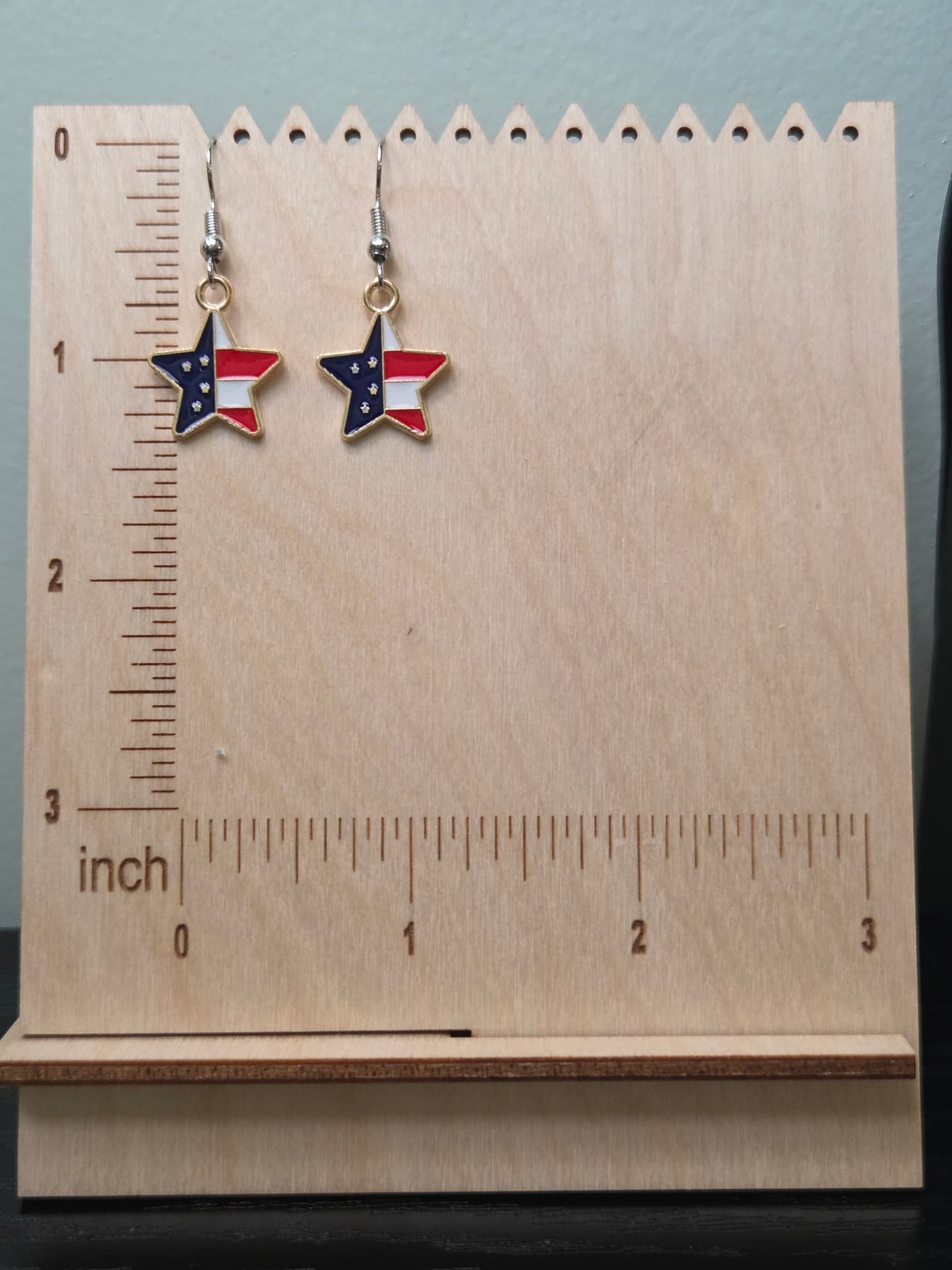 Pair of petite American flag split star dangle earrings on a wooden display board with ruler showing approximately ½ inch charm size, featuring navy blue star upper left and red and white stripe lower right in a gold-tone frame