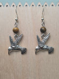 Hummingbird charm and tiger's eye gemstone earrings resting on a natural wood panel, highlighting the antiqued silver-tone finish and earthy stone