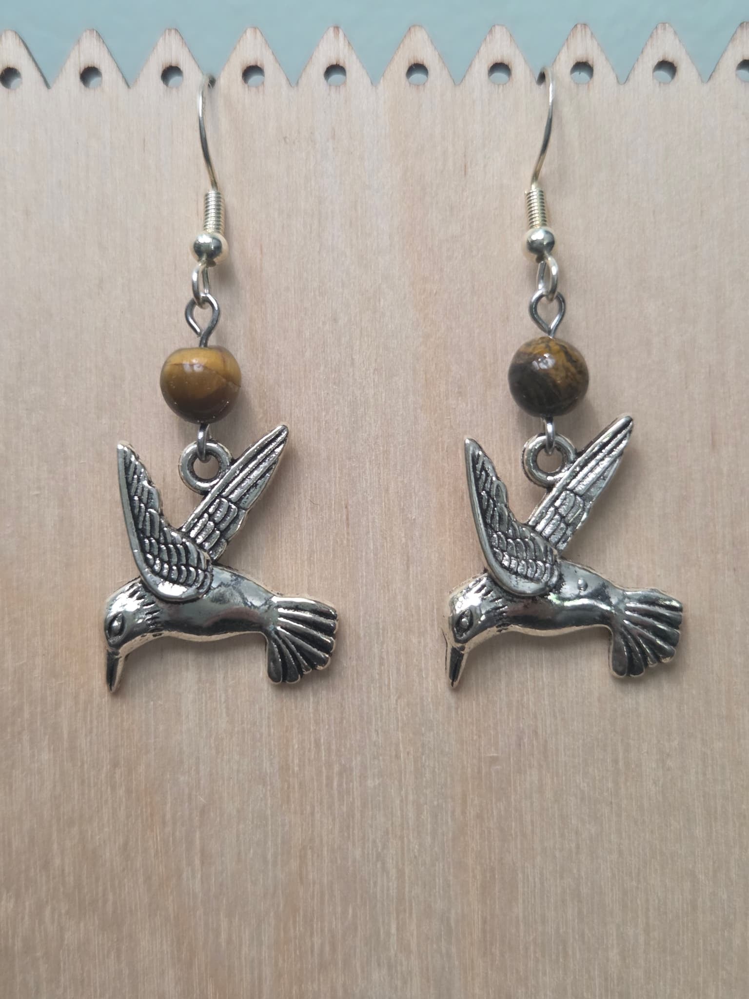 Hummingbird charm and tiger's eye gemstone earrings resting on a natural wood panel, highlighting the antiqued silver-tone finish and earthy stone