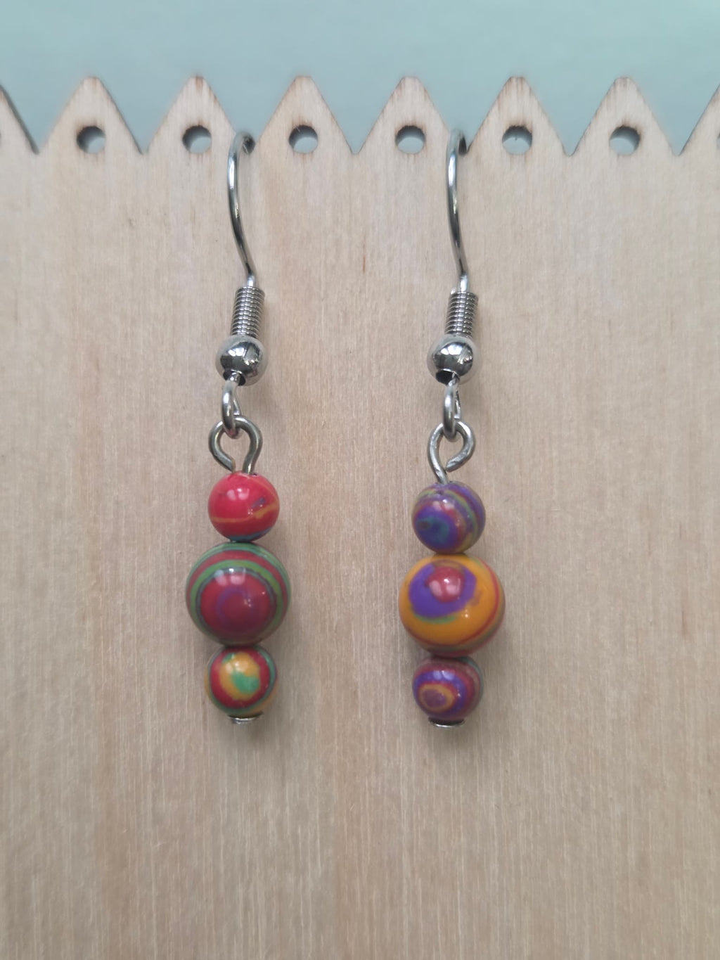 Handmade graduated drop earrings featuring three round mystery stone beads swirled in rich jewel tones of red, green, purple, gold, and orange — two mismatched earrings shown side by side on a wood display