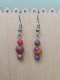 Handmade graduated drop earrings featuring three round mystery stone beads swirled in rich jewel tones of red, green, purple, gold, and orange — two mismatched earrings shown side by side on a wood display