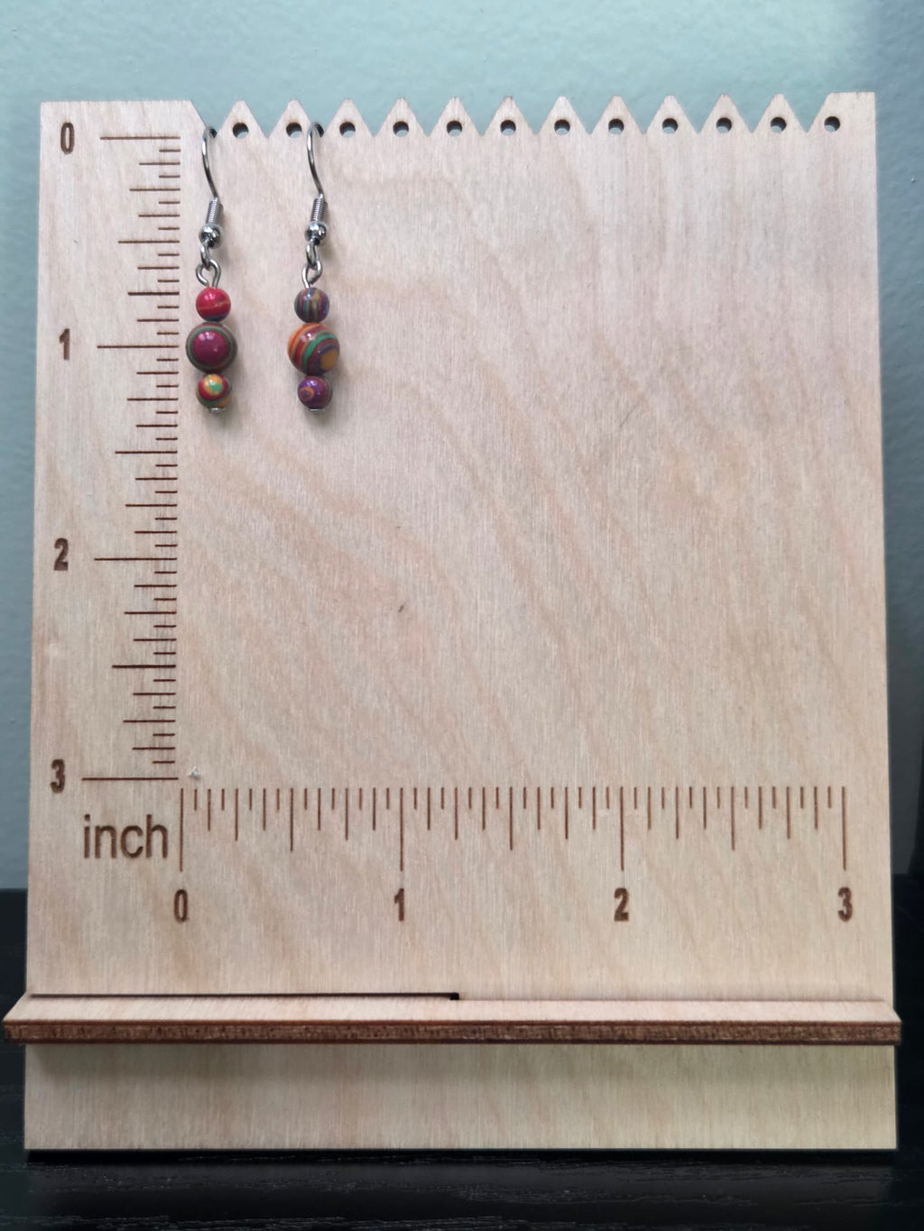 Jewel tone mystery stone graduated drop earrings on a ruler display — approximately 1.5 inches long — showing vibrant swirled bead detail in red, green, purple, gold, and orange