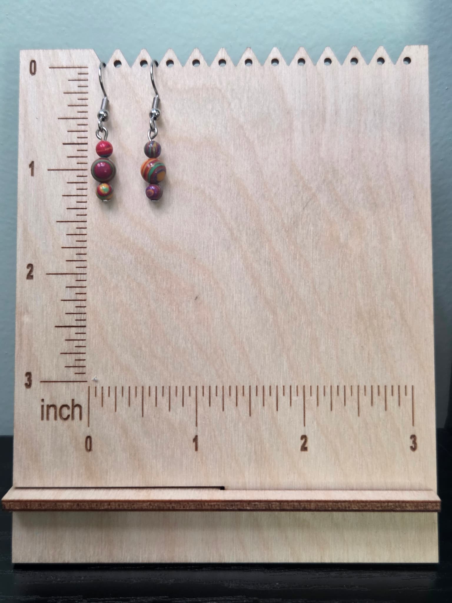 Jewel tone mystery stone graduated drop earrings on a ruler display — approximately 1.5 inches long — showing vibrant swirled bead detail in red, green, purple, gold, and orange