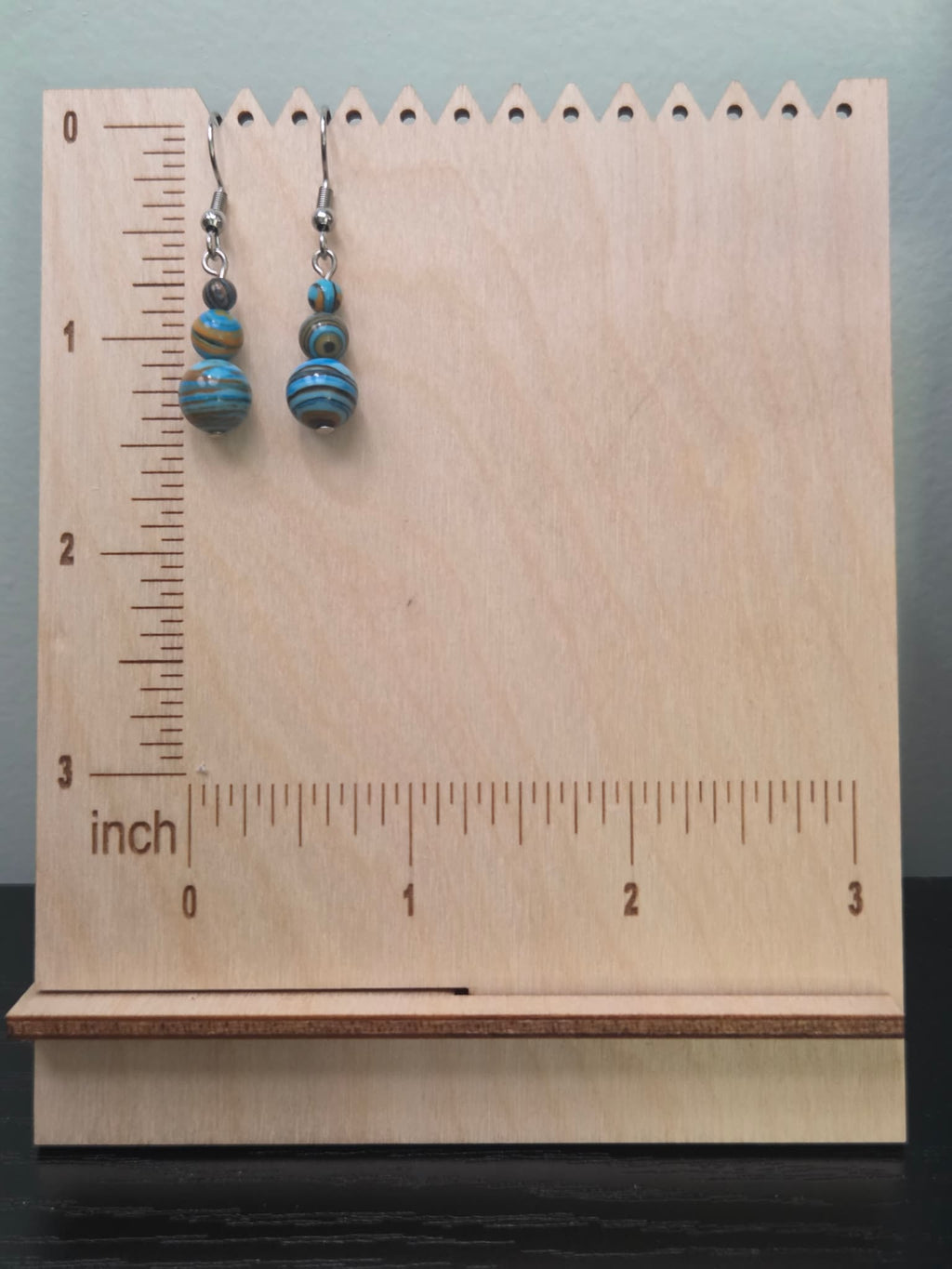 Mystery stone graduated drop earrings shown on a ruler display — approximately 1.5 inches long — in blue, amber, and brown swirl beads with stainless steel ear wires