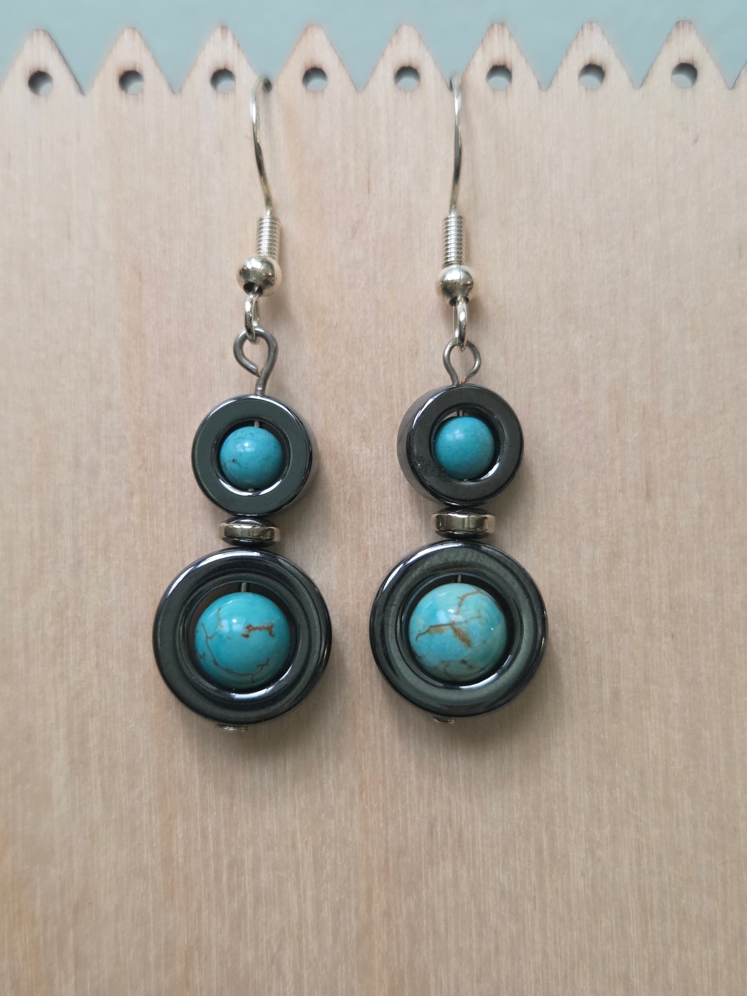 Hematite and turquoise howlite halo drop earrings laid flat on a wooden surface, showing two tiers of round turquoise stones set in gunmetal hematite rings connected by a silver spacer